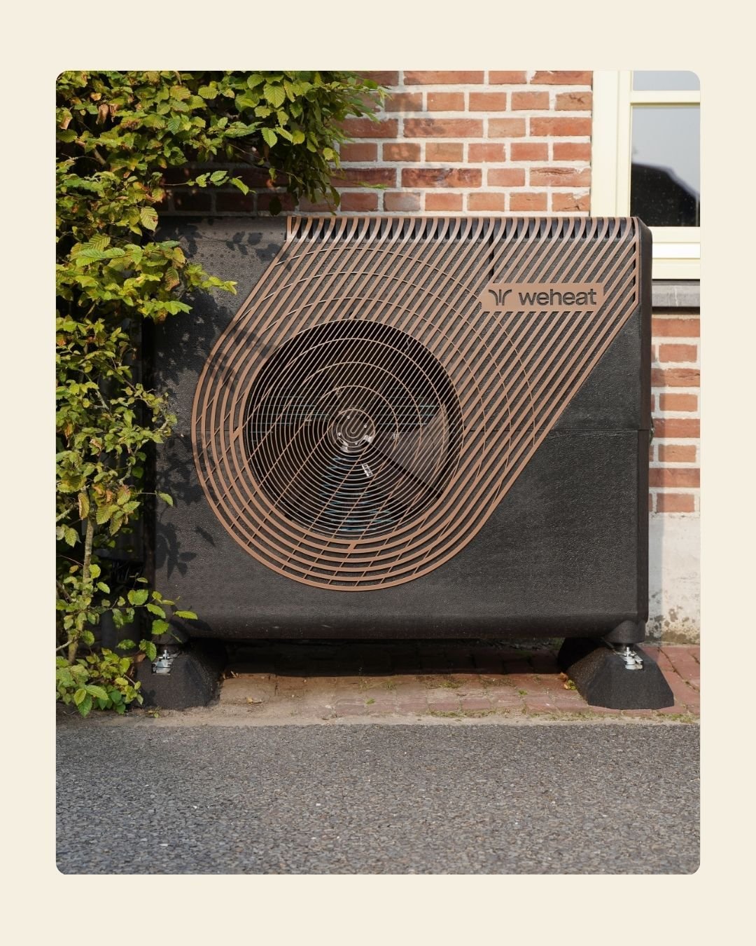 Een buitenlucht airconditioner van het merk WeHeat, geplaatst naast een bakstenen muur en een raam, met groen struikgewas links. Buitenunit warmtepomp Sparrow