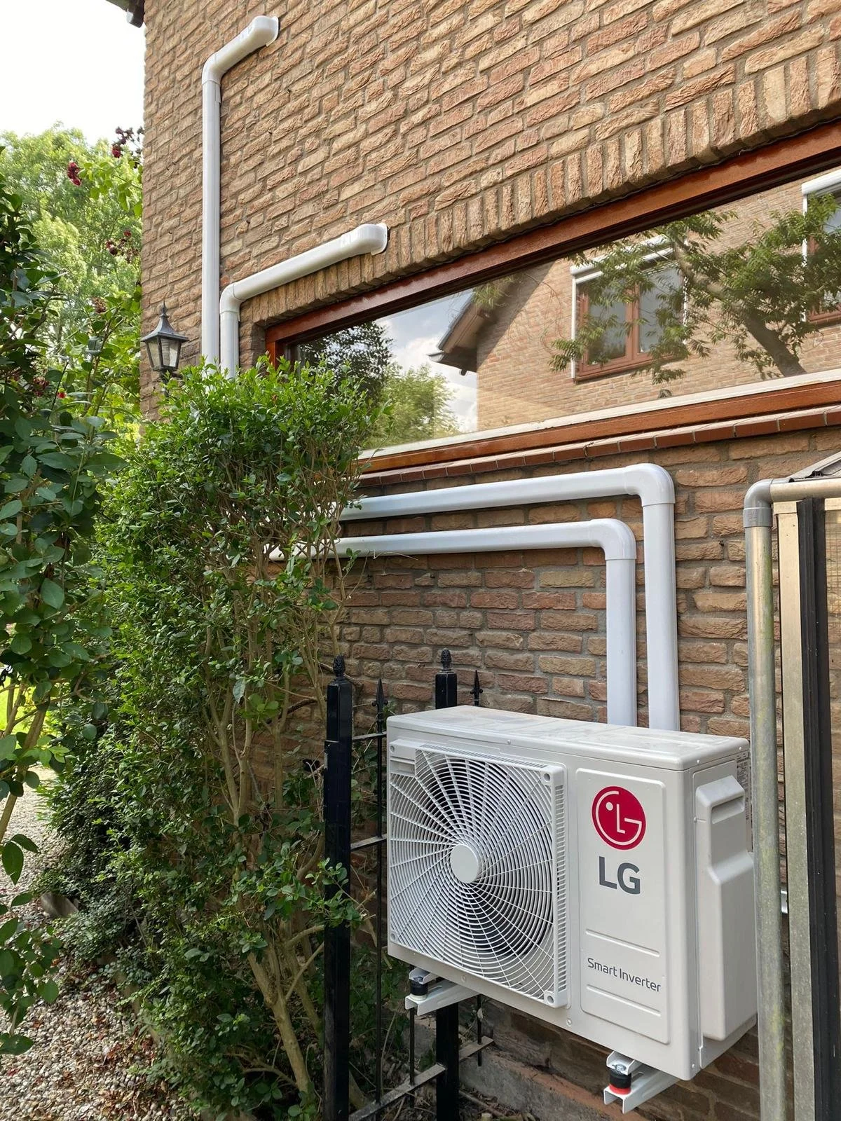 De afbeelding toont een buitenmuur van een huis met een raam, een klimopplant en een buitenunit van een airconditioner van LG. Er zijn ook witte PVC-buizen bevestigd aan de muur.