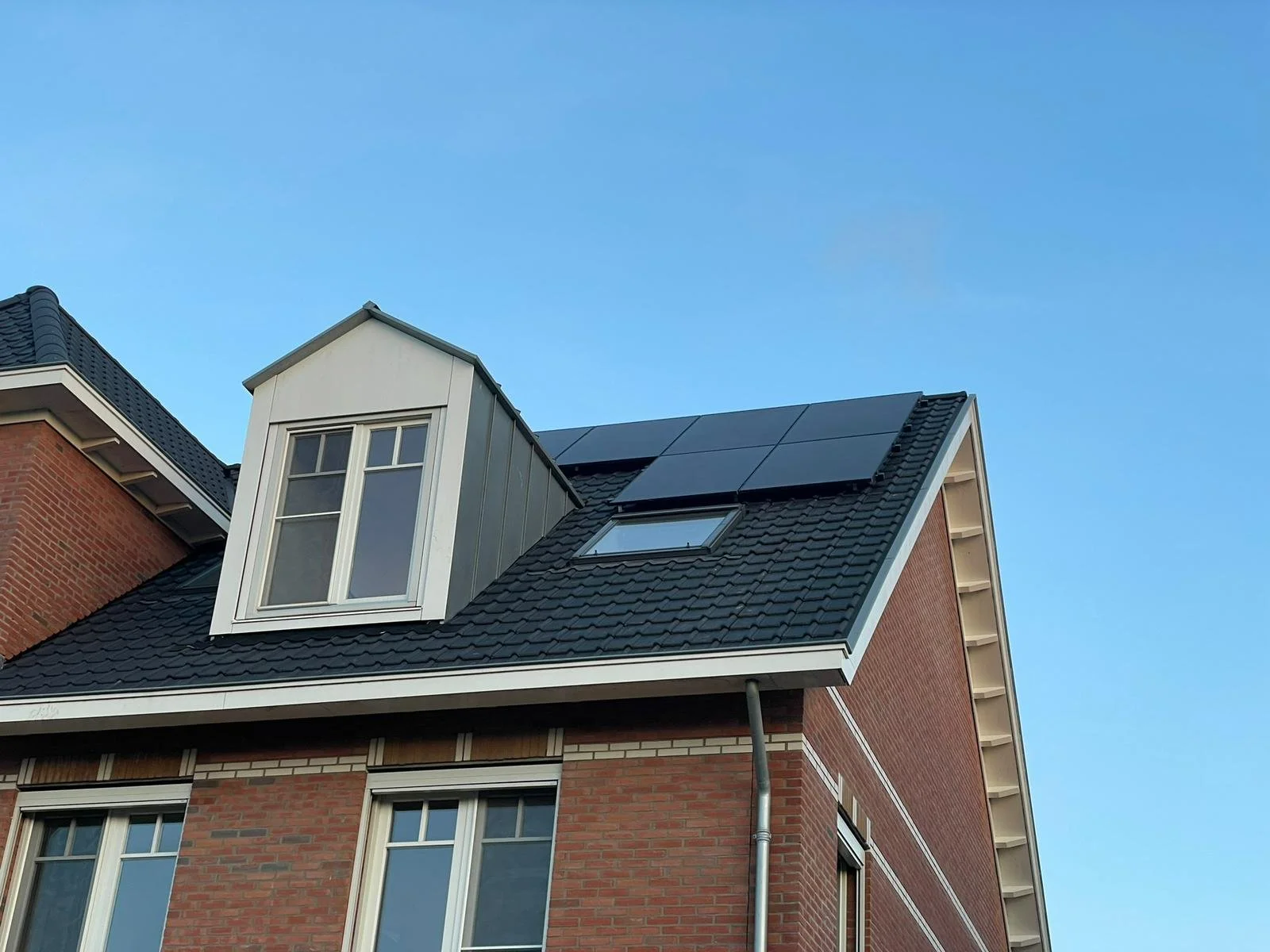 Daken van een modern huis met zonnepanelen en een dakkapel onder een heldere blauwe hemel.