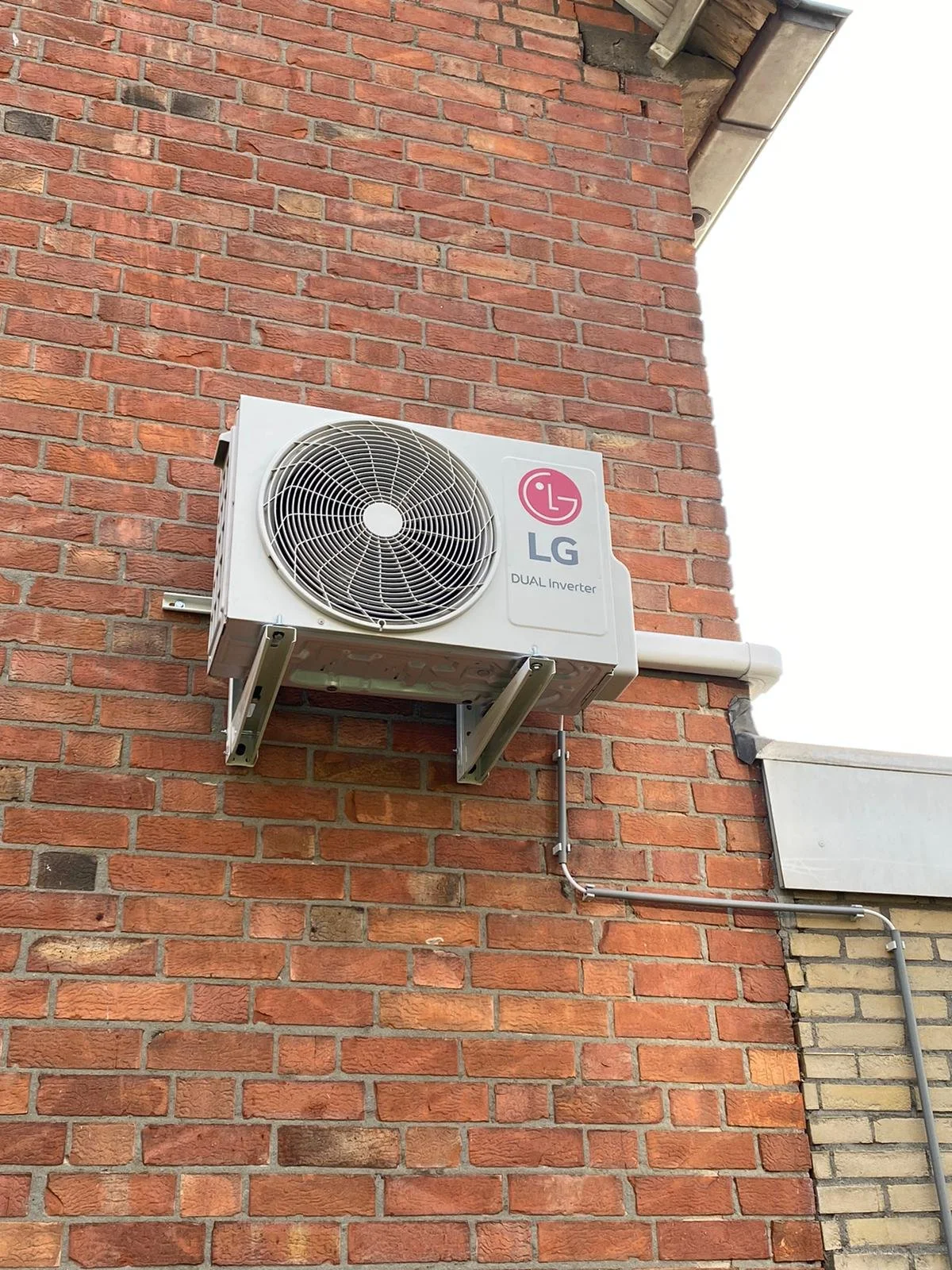 Buitenmuur van een gebouw met een airconditioningsunit van LG, gemonteerd op een metalen steun aan een bakstenen muur.