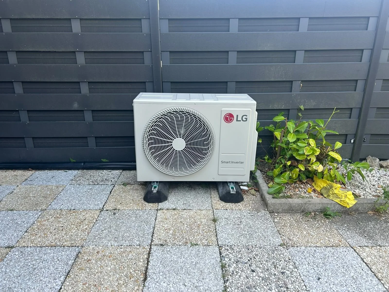 Buiten airconditioningunit van LG naast een plantenbak met groen blad en een gele blad, met een zwart houten hek op de achtergrond.