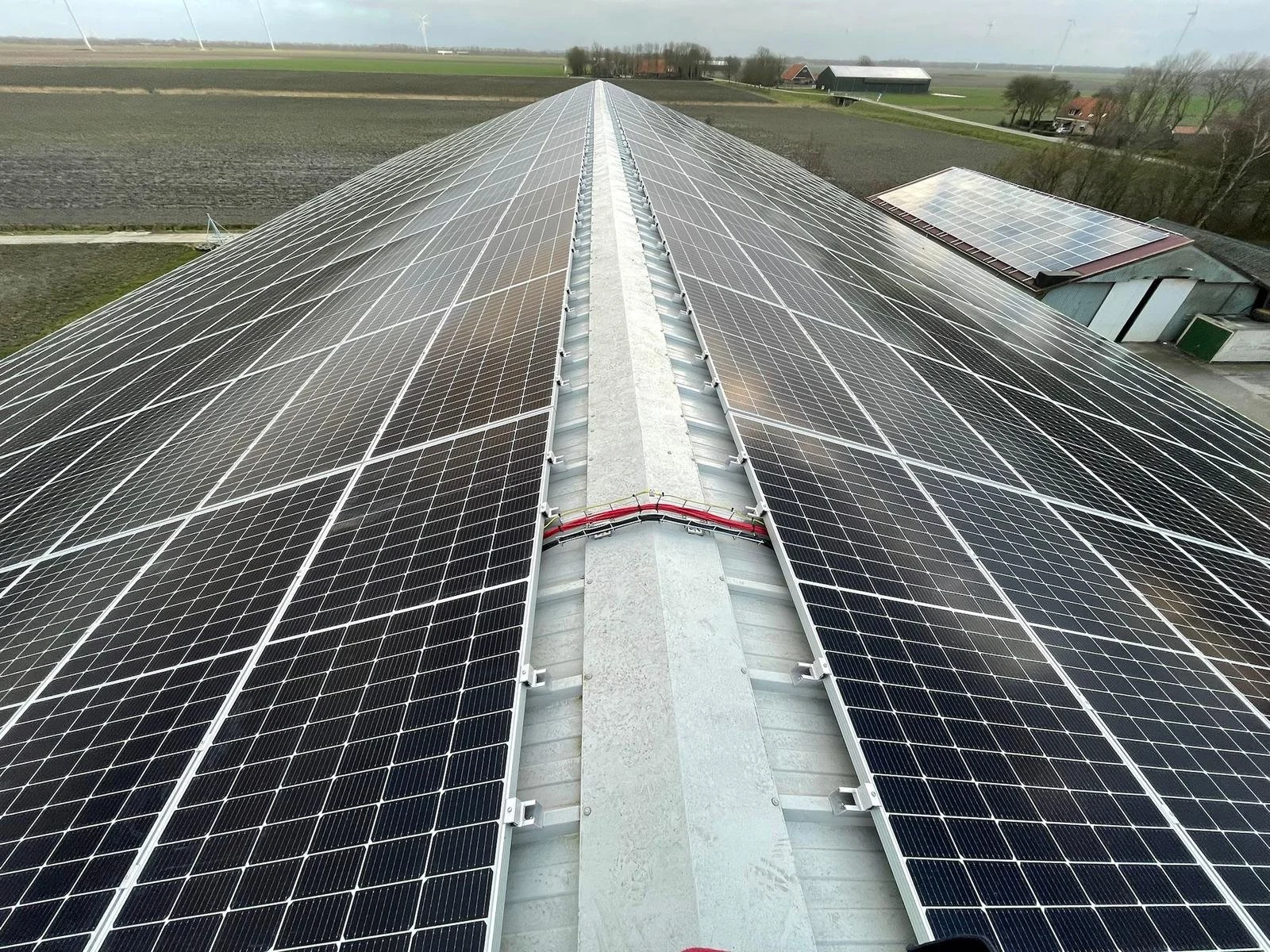 Zonnedak met zonnepanelen op platteland, omringd door velden en bomen, met windturbines op de achtergrond.