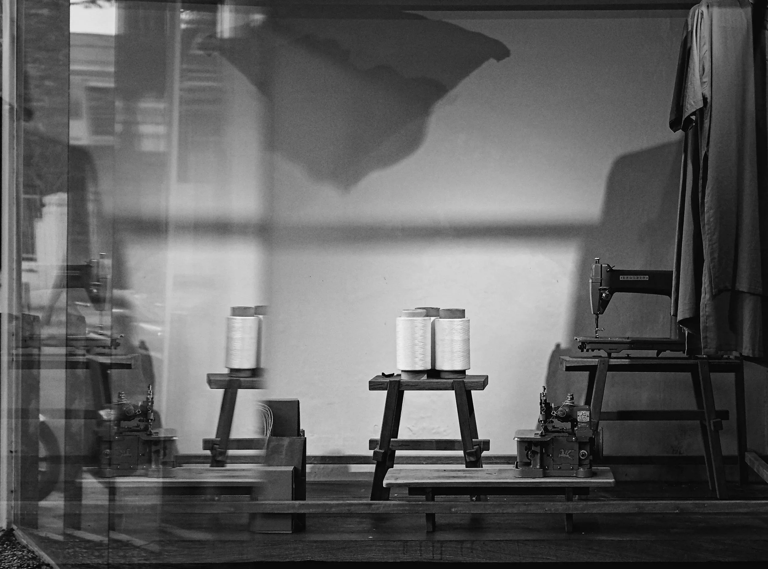 A black and white photo of a sewing workspace. The foreground has three tall spools of thread placed on a small table. In the middle, there's a larger table with fabric or other materials. A sewing machine labeled "Brother" is visible on the right side, with some clothing hanging beside it. The background appears to be a wall with factory or store shelves reflected on a glass, and the overall scene has a vintage or industrial feel.