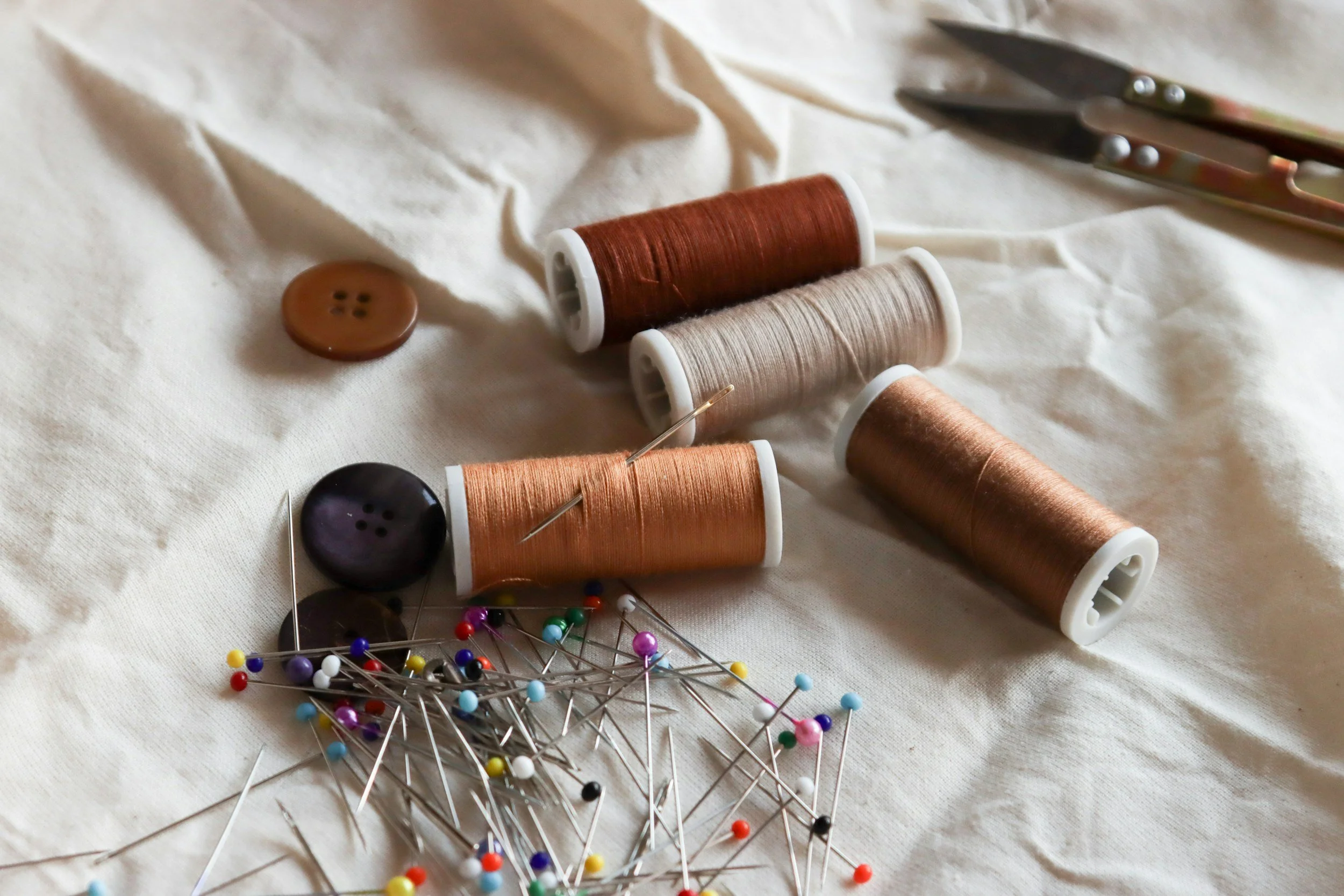 Various sewing supplies including spools of thread in shades of brown and beige, two buttons, straight pins with colored heads, and a pair of scissors on a white fabric surface.