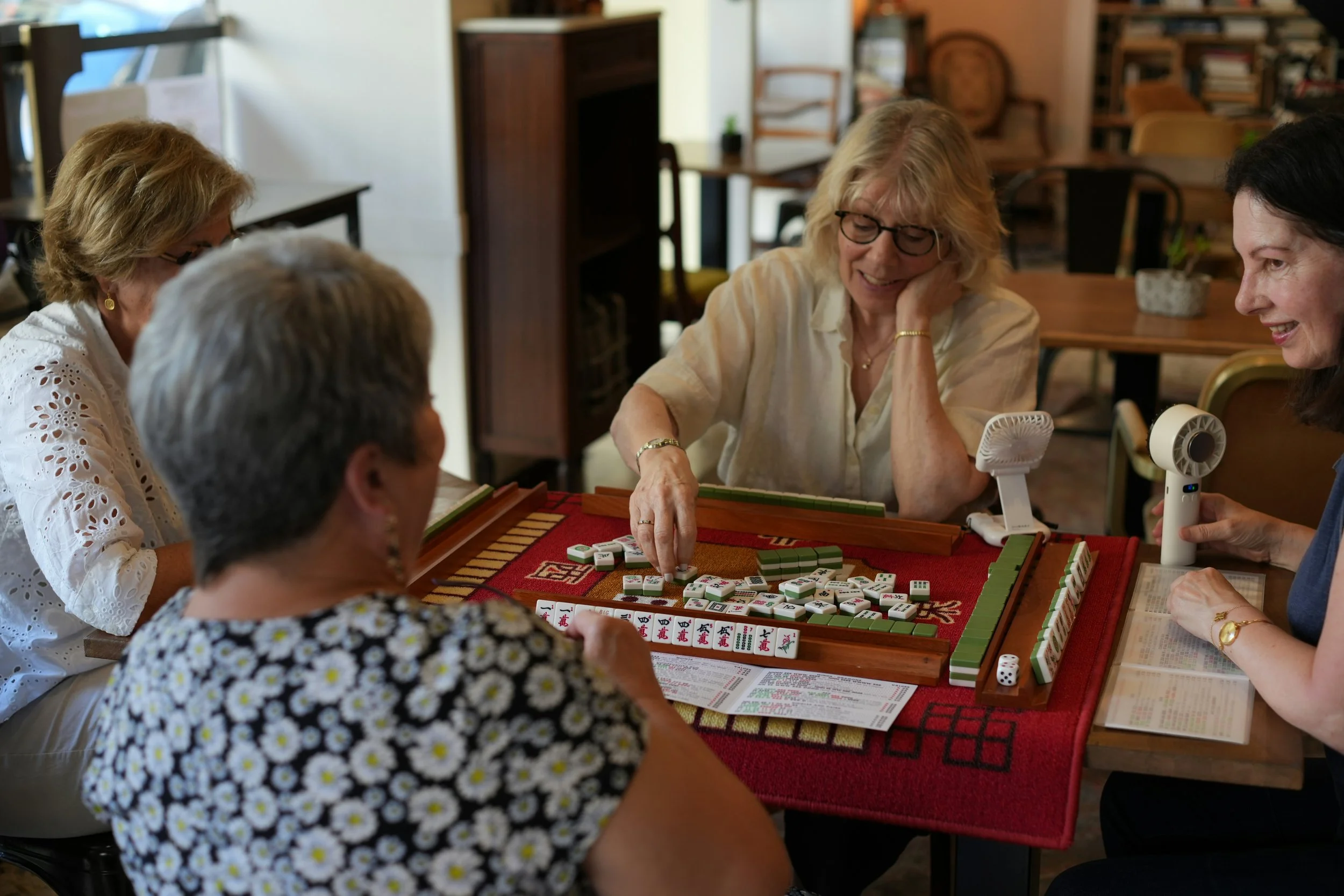 Four women gathered around a table playing American Mahjong in a comfortable indoor setting with bookshelves and wooden furniture, representing the welcoming community experience of Mahjong Awaits™.