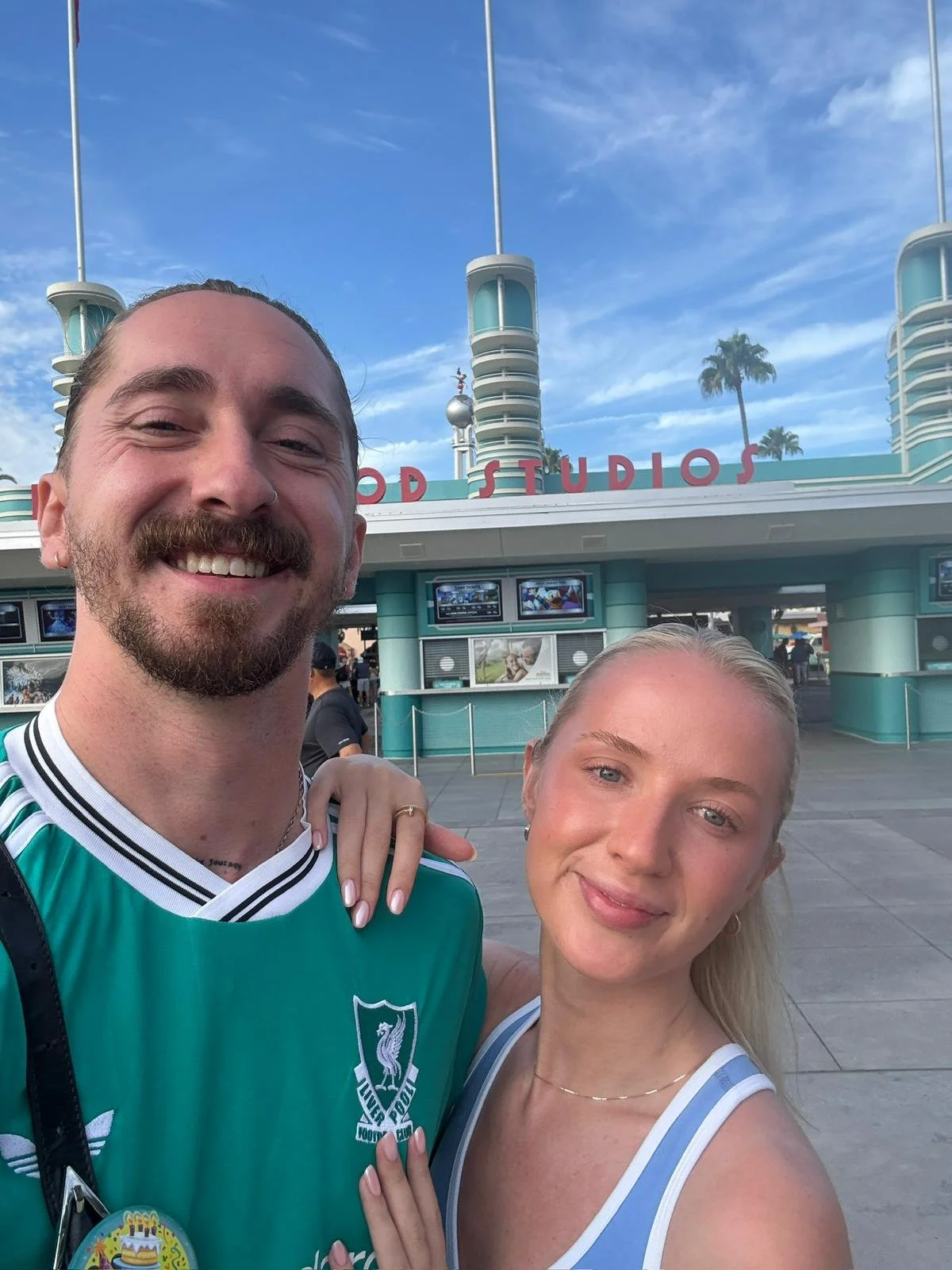 A smiling man and woman taking a selfie in front of the entrance to a Disney theme park, with the sign 'Walt Disney Studios' visible in the background.