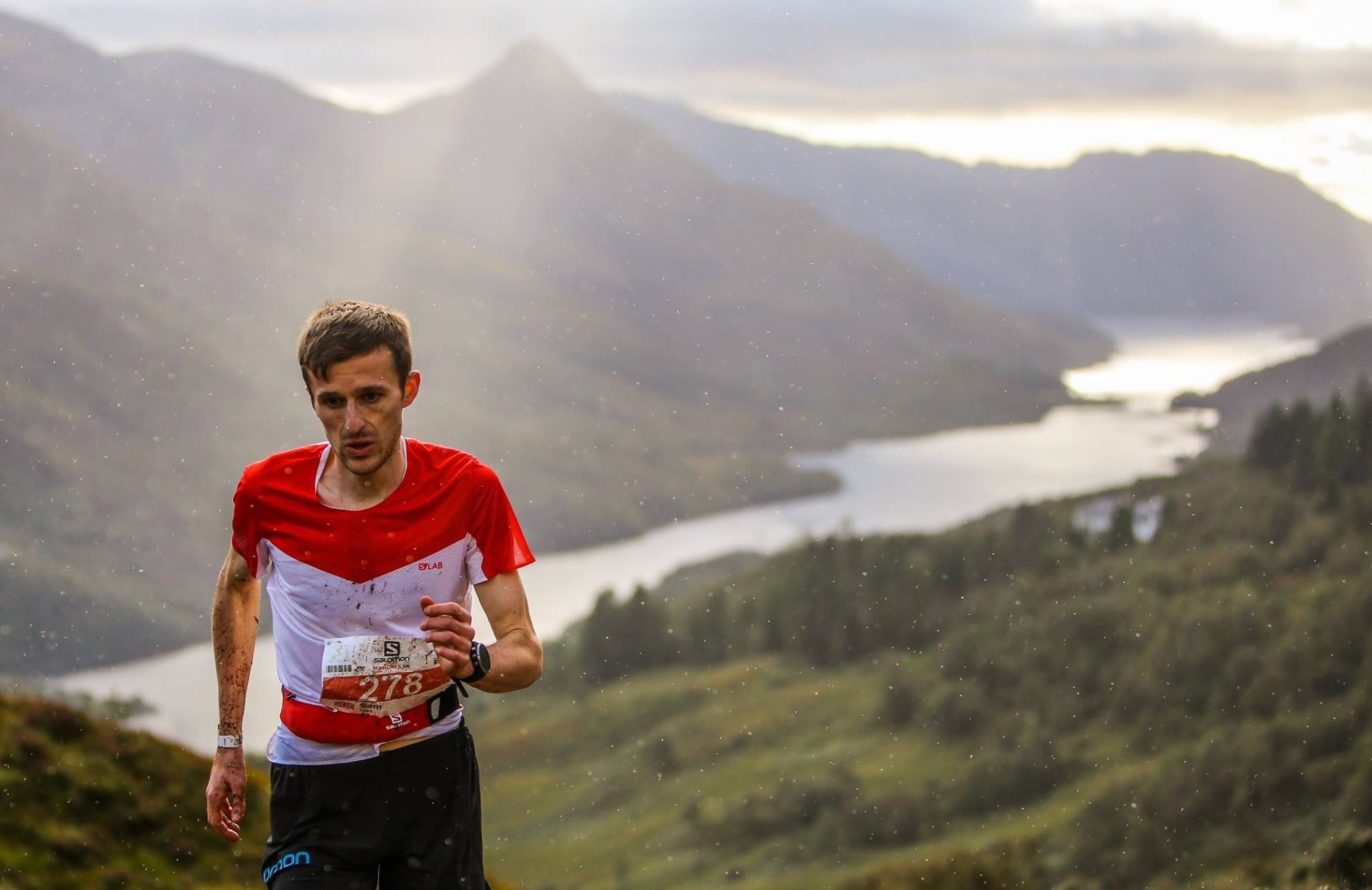 A male runner, wearing a white and red shirt with a race bib, running outdoors in a scenic area with mountains and a lake in the background, during what appears to be a race.