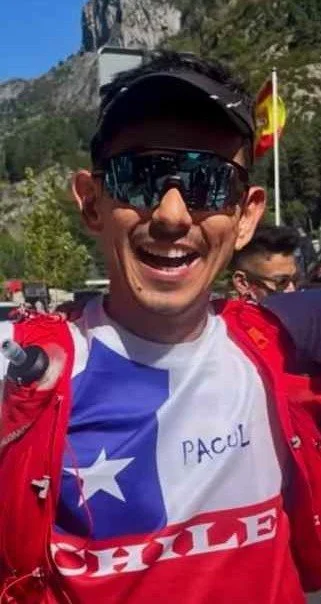 Smiling man with sunglasses, wearing a Chilean flag-themed shirt and red jacket, outdoors with a mountain and Chilean flag in the background.