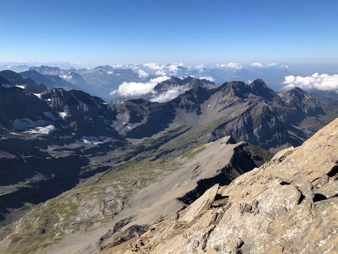 Mountain landscape with rugged peaks, some patches of snow, green valleys, and clouds in the sky.