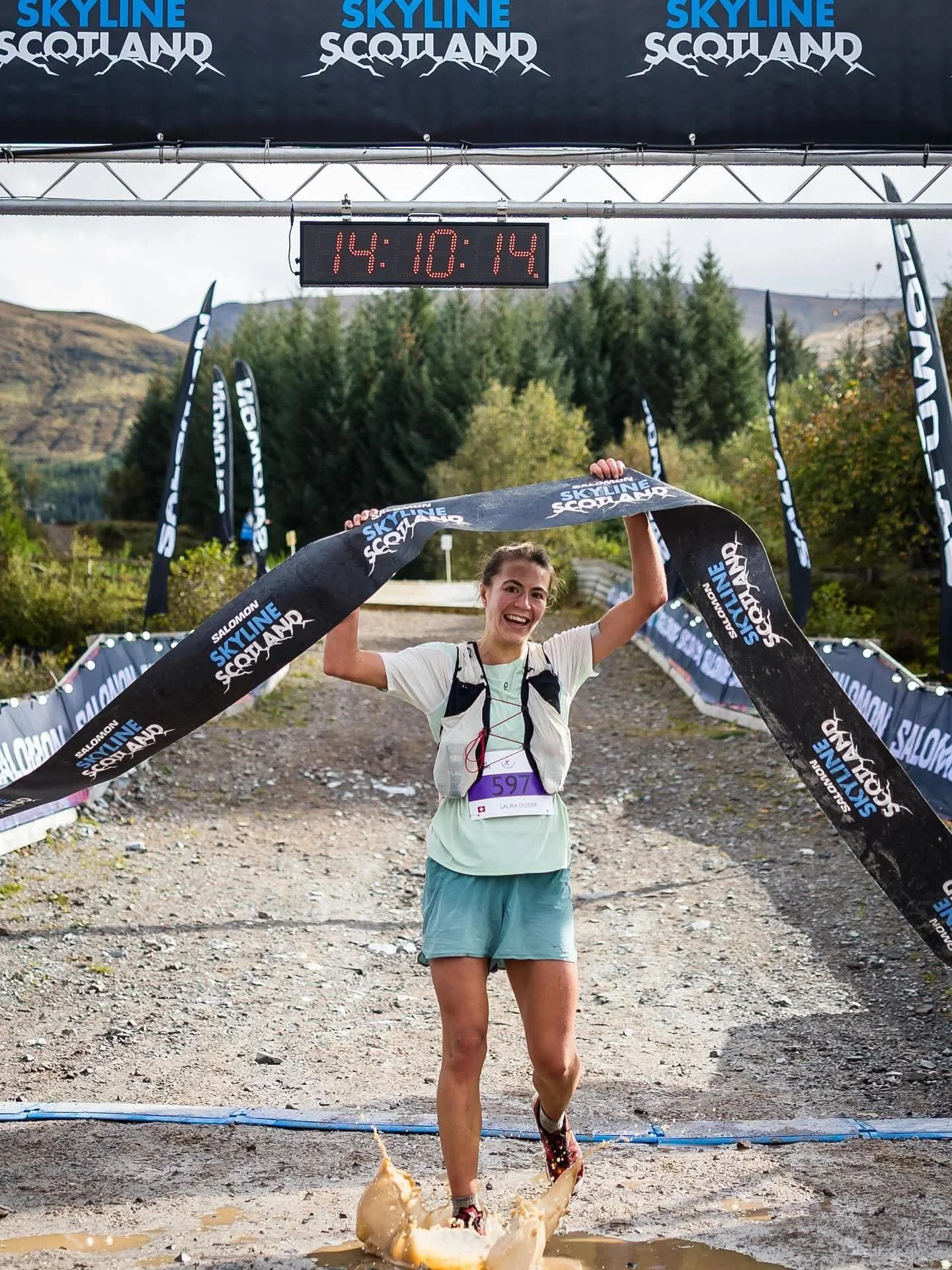A woman is smiling and holding a race tape at the finish line of an outdoor trail race with trees and hills in the background. She is wearing a light-colored shirt, teal shorts, and running shoes, and is crossing through a muddy puddle.