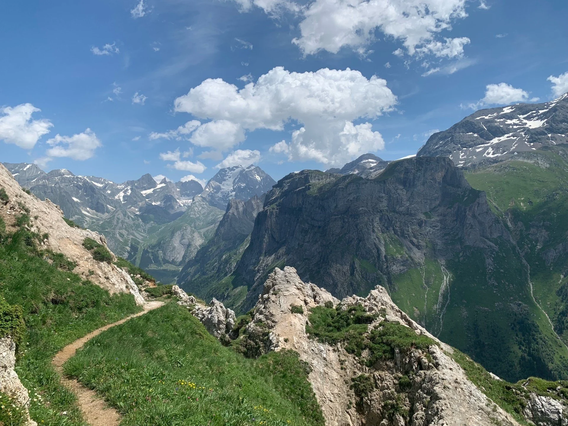 A mountain trail winds through green grass and rocks, with rugged snow-capped mountains and a bright blue sky with scattered clouds in the background.