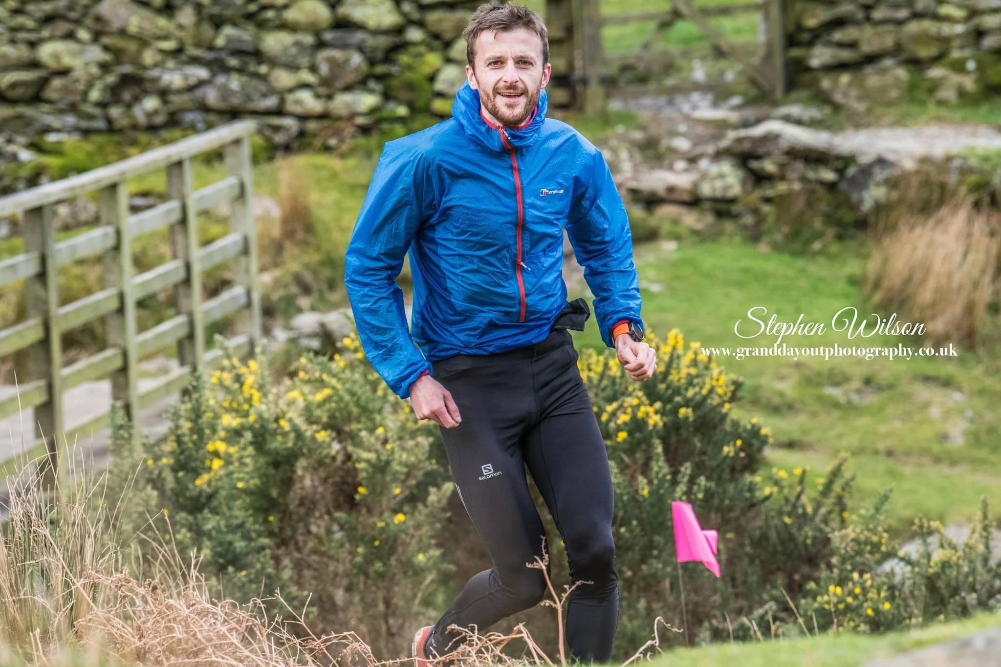 A man in a blue waterproof jacket and black athletic pants running outdoors on a trail in a grassy area with yellow flowers, stone walls, and fences in the background.