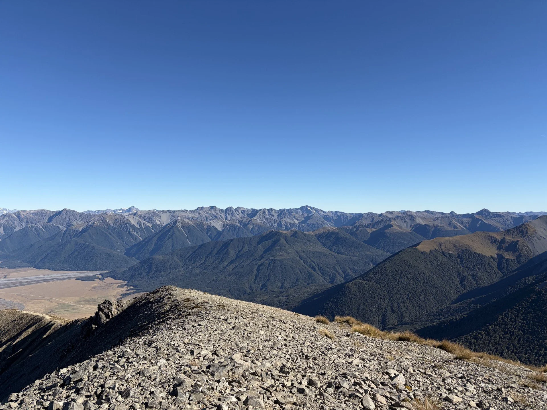 A wide view of rugged mountain range under clear blue sky with no clouds, featuring multiple peaks with some snow caps, and a rocky foreground area.