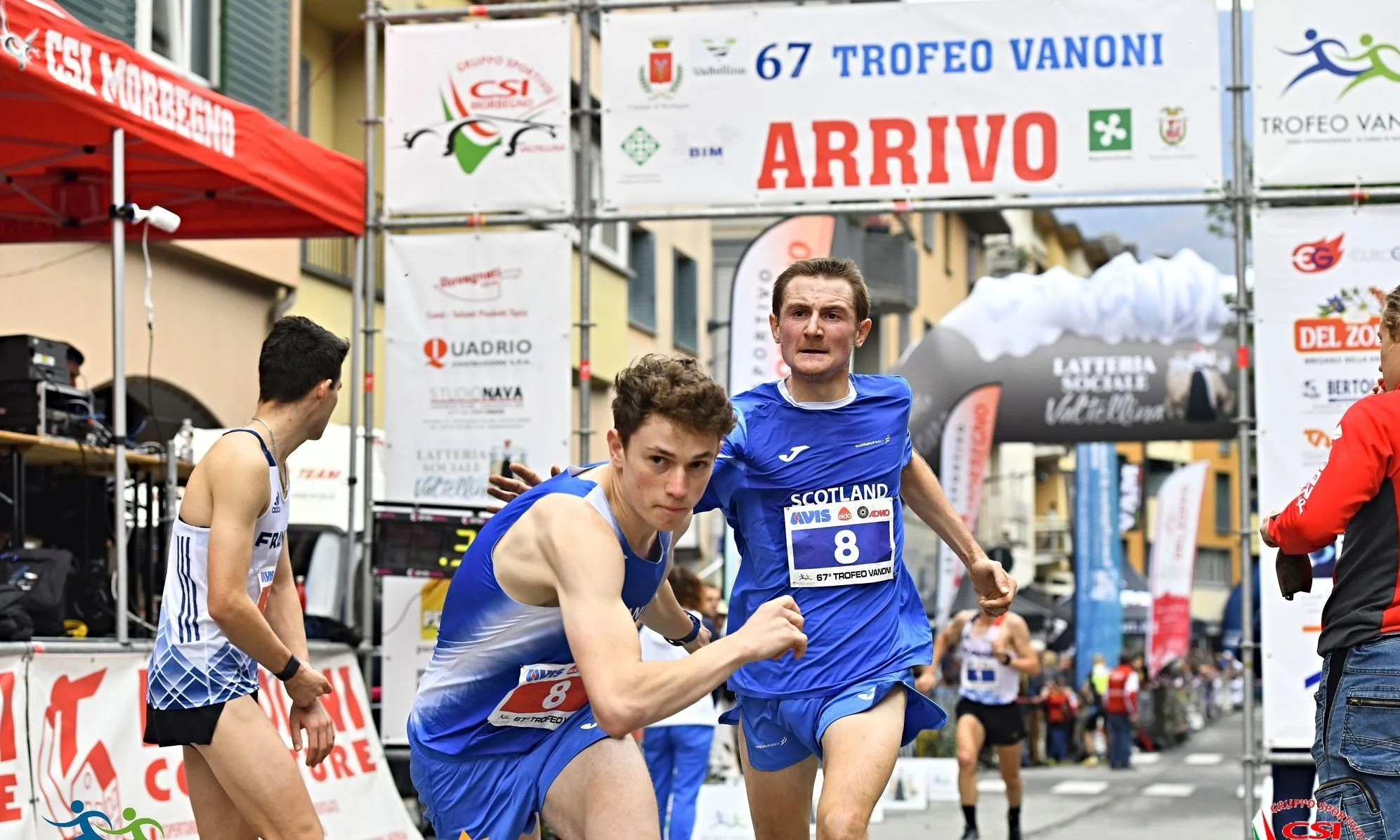 Male runners in a street race, wearing blue and white athletic gear, nearing the finish line marked 'ARRIVO' with a banner overhead, surrounded by race officials and spectators.