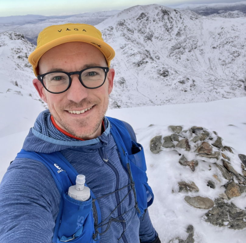 A man taking a selfie in a snowy mountain landscape, wearing a yellow cap, glasses, a blue jacket, and carrying a hydration pack.