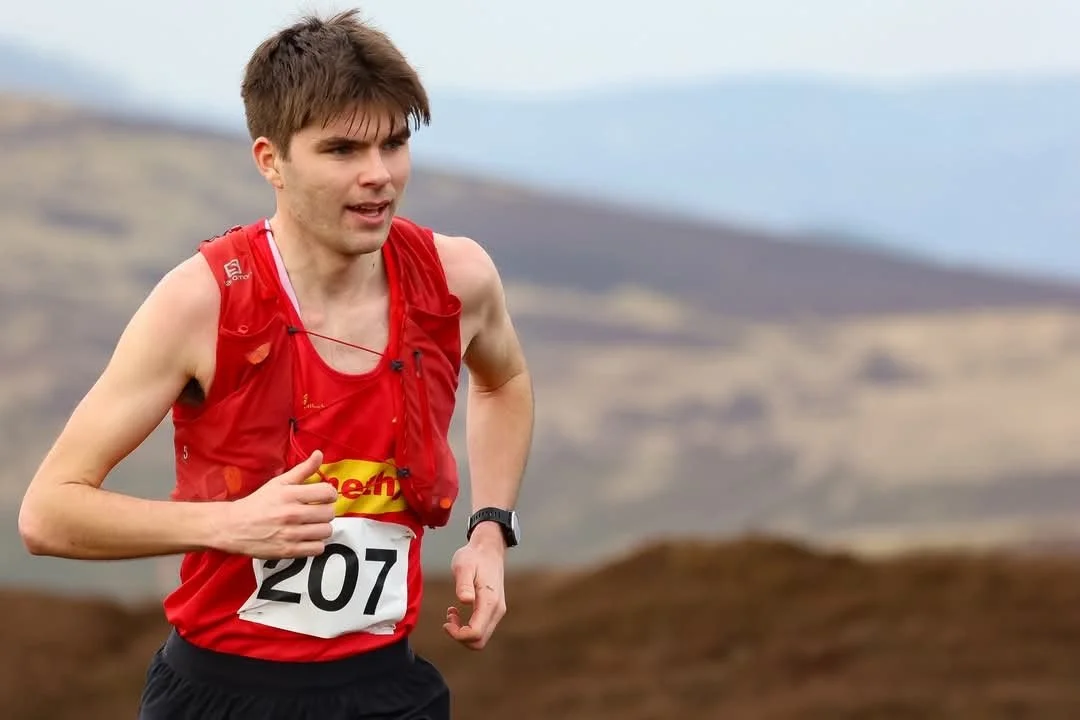 A young man running outdoors during a race, wearing a red athletic tank top with the number 207, and a hydration pack.