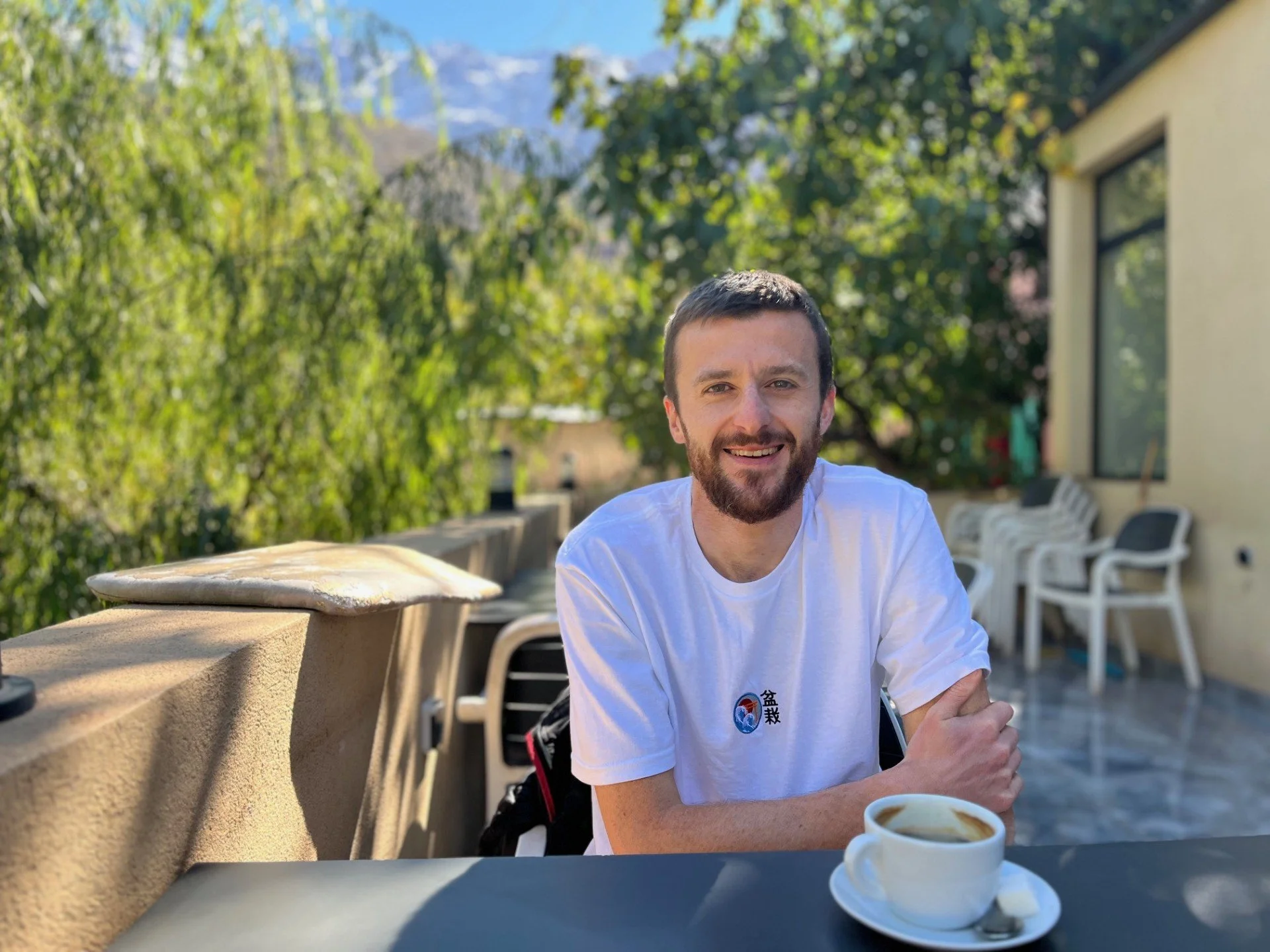 A man with a beard and short brown hair smiling at the camera while sitting at a table outdoors, with trees and mountains in the background. He is wearing a white t-shirt with a logo on it, and there is a cup of coffee or tea in front of him.