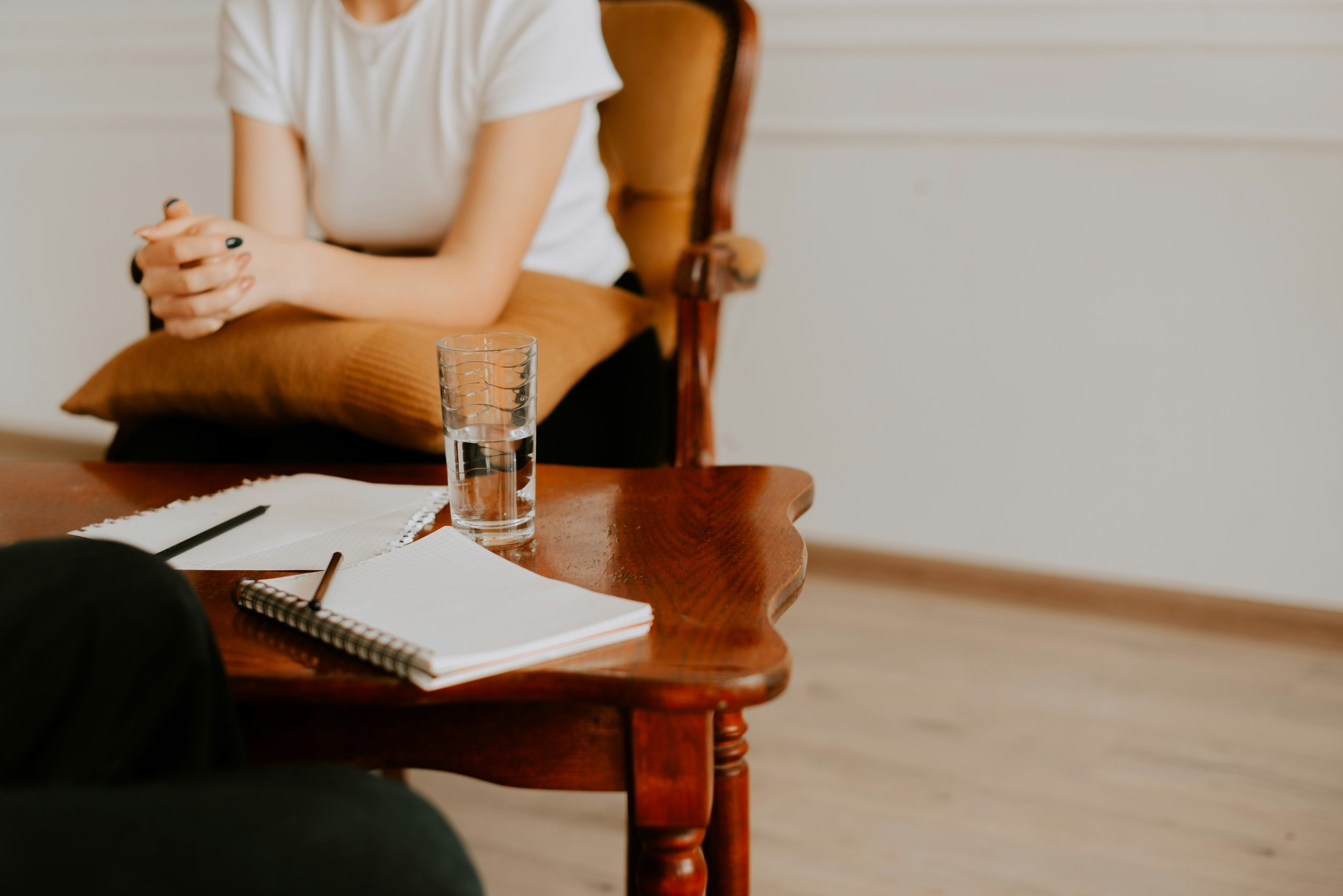 Person sitting at a wooden table with a cup of water, notebook, and pen, with hands clasped.