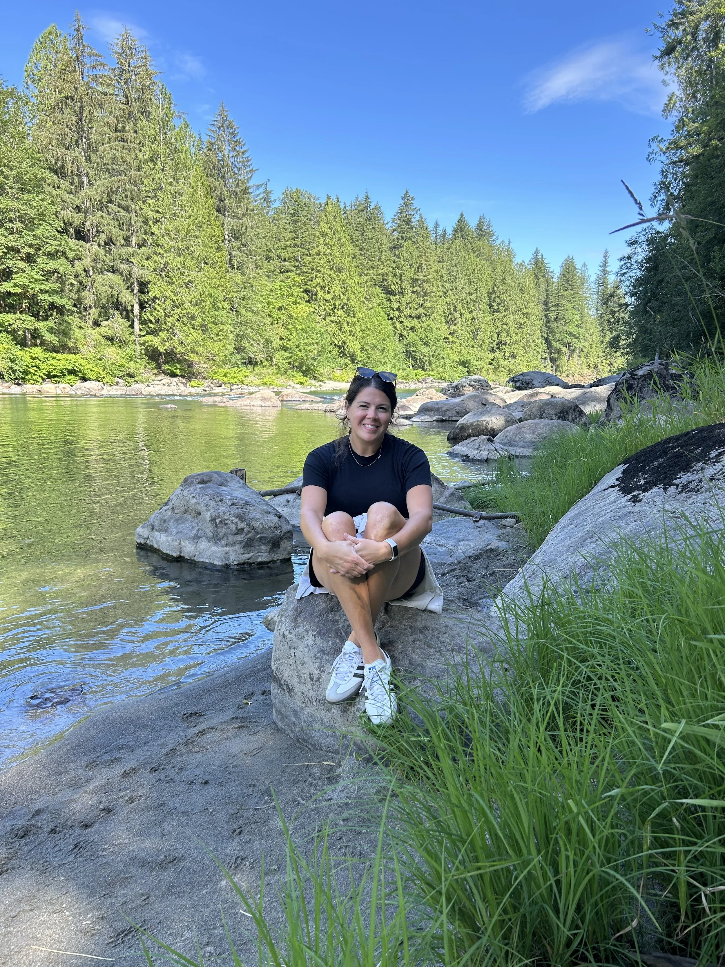 A smiling woman sitting on a large rock by a river in a forest with tall trees and green foliage under a blue sky.
