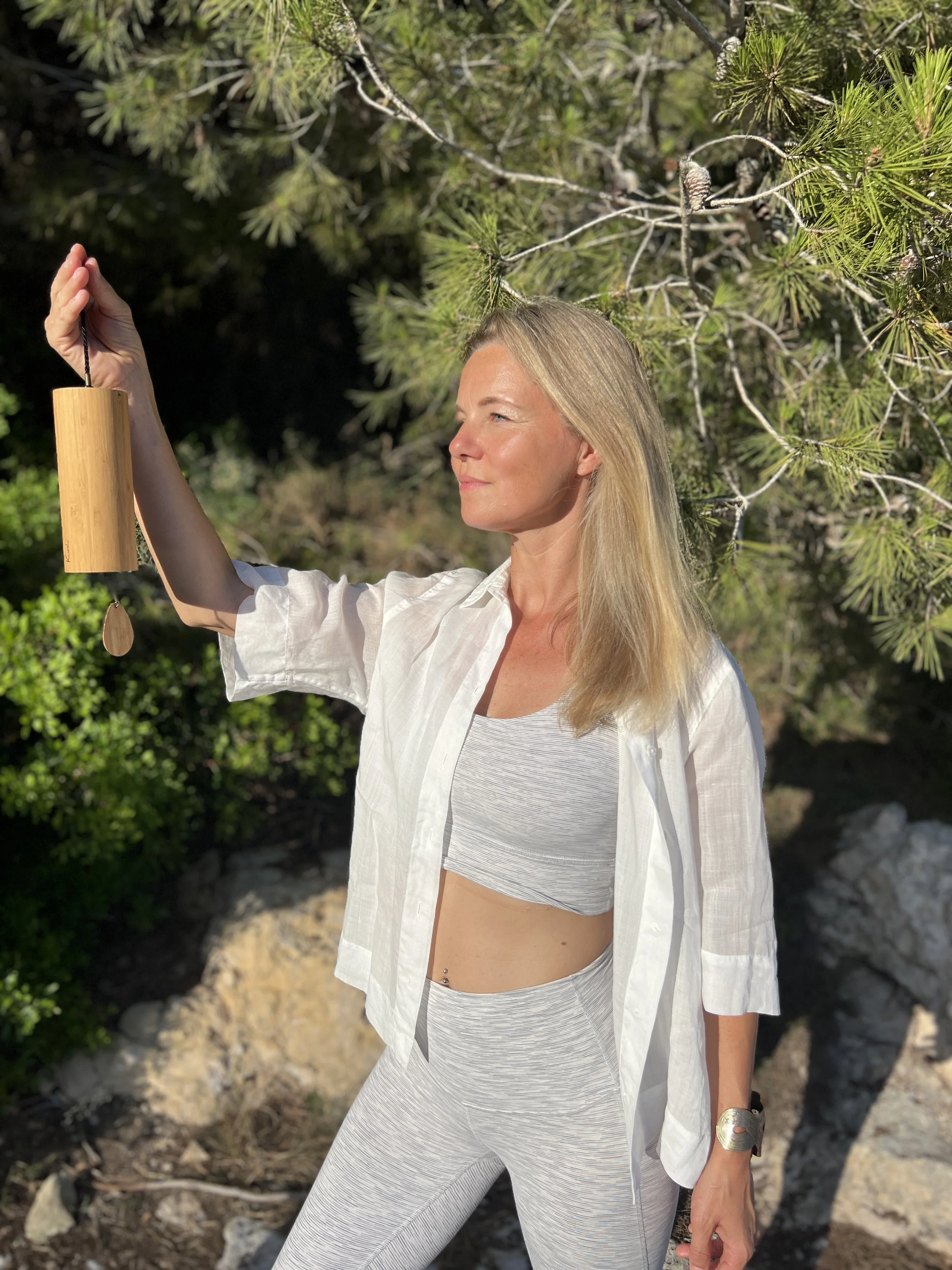 A woman with blonde hair wearing a white open shirt over a sports bra and leggings, standing outdoors near trees, holding a wooden wind chime.