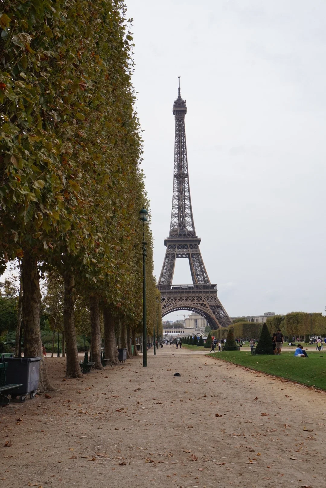 Eiffel Tower in Paris, viewed from a nearby park with trees lining a pathway, people walking and sitting on the grass, mostly overcast sky.