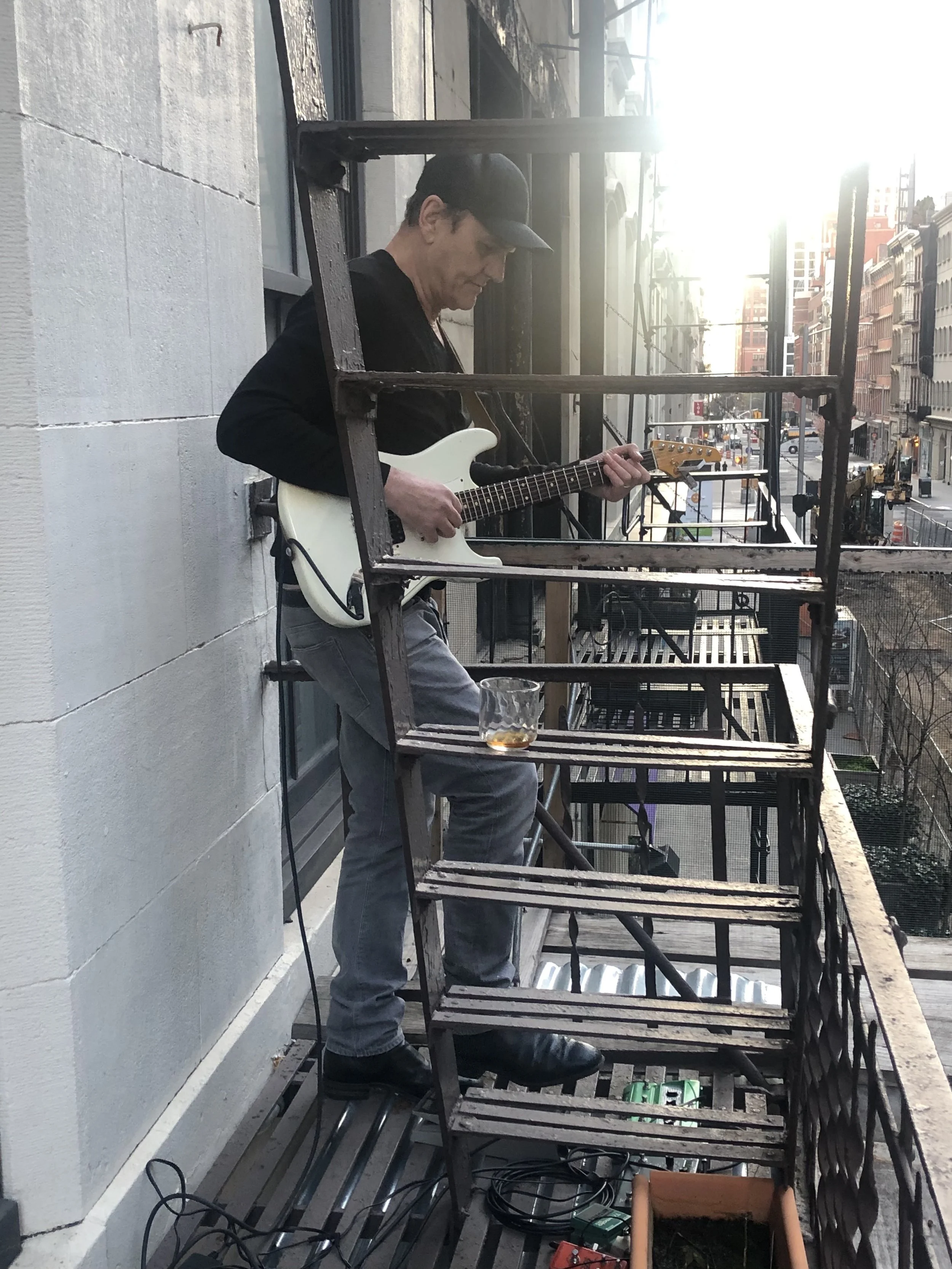 A man wearing a black baseball cap and gray pants playing an electric guitar on a small balcony in sunlight, with urban buildings and street visible in the background.