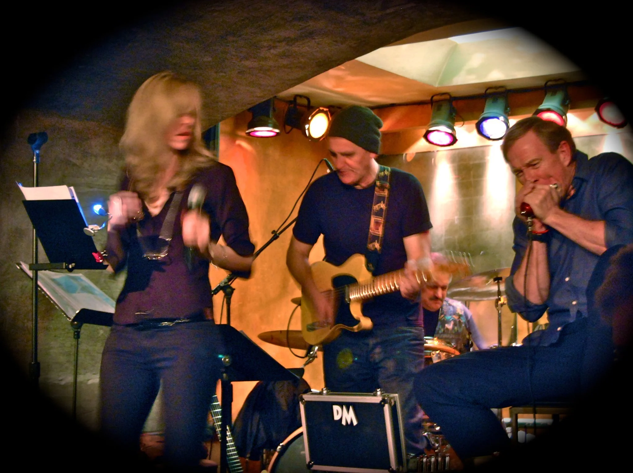 Three musicians performing on stage in a dimly lit venue. One woman with blonde hair holds a tambourine, a man wearing a beanie plays an acoustic guitar, and another man is singing into a microphone. Musical equipment and stage lights are visible.