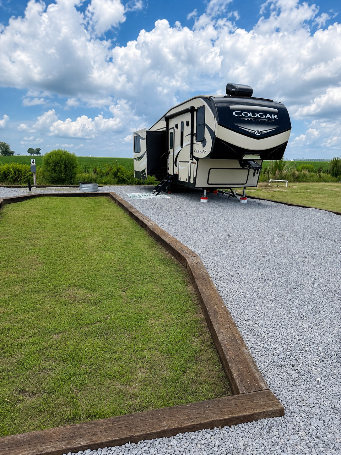 Travel trailer parked on a gravel pad next to a small grassy area, with open fields and a partly cloudy sky in the background.