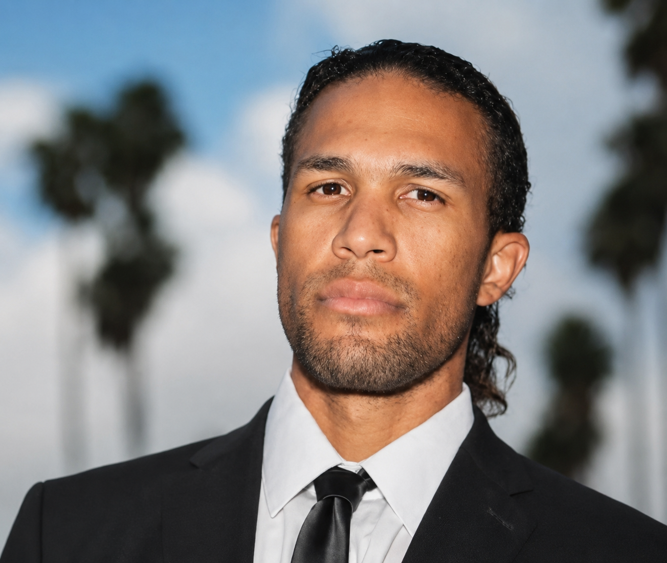 A man in a black tuxedo with a white shirt and black tie, standing outdoors with palm trees and a cloudy sky in the background.