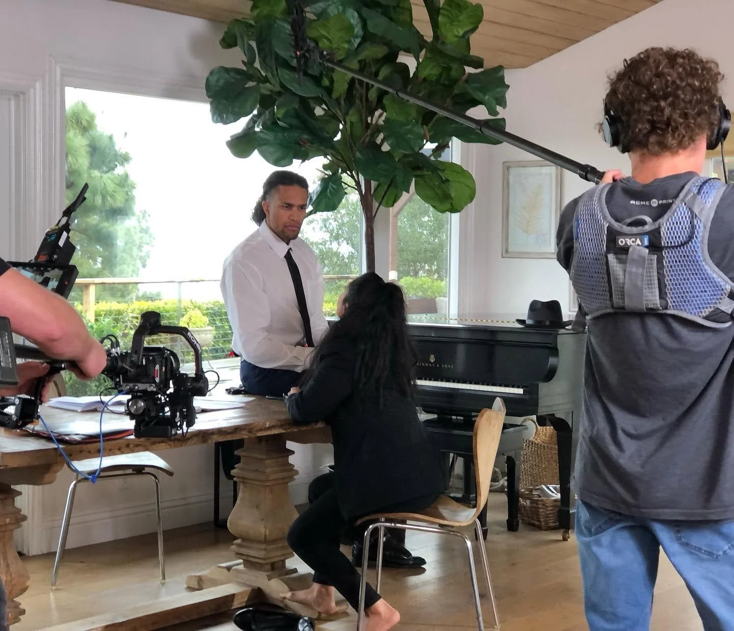 Filming scene inside a house, featuring a man in a white shirt and black tie talking to a woman at a grand piano, with a film crew and cameras capturing the moment.