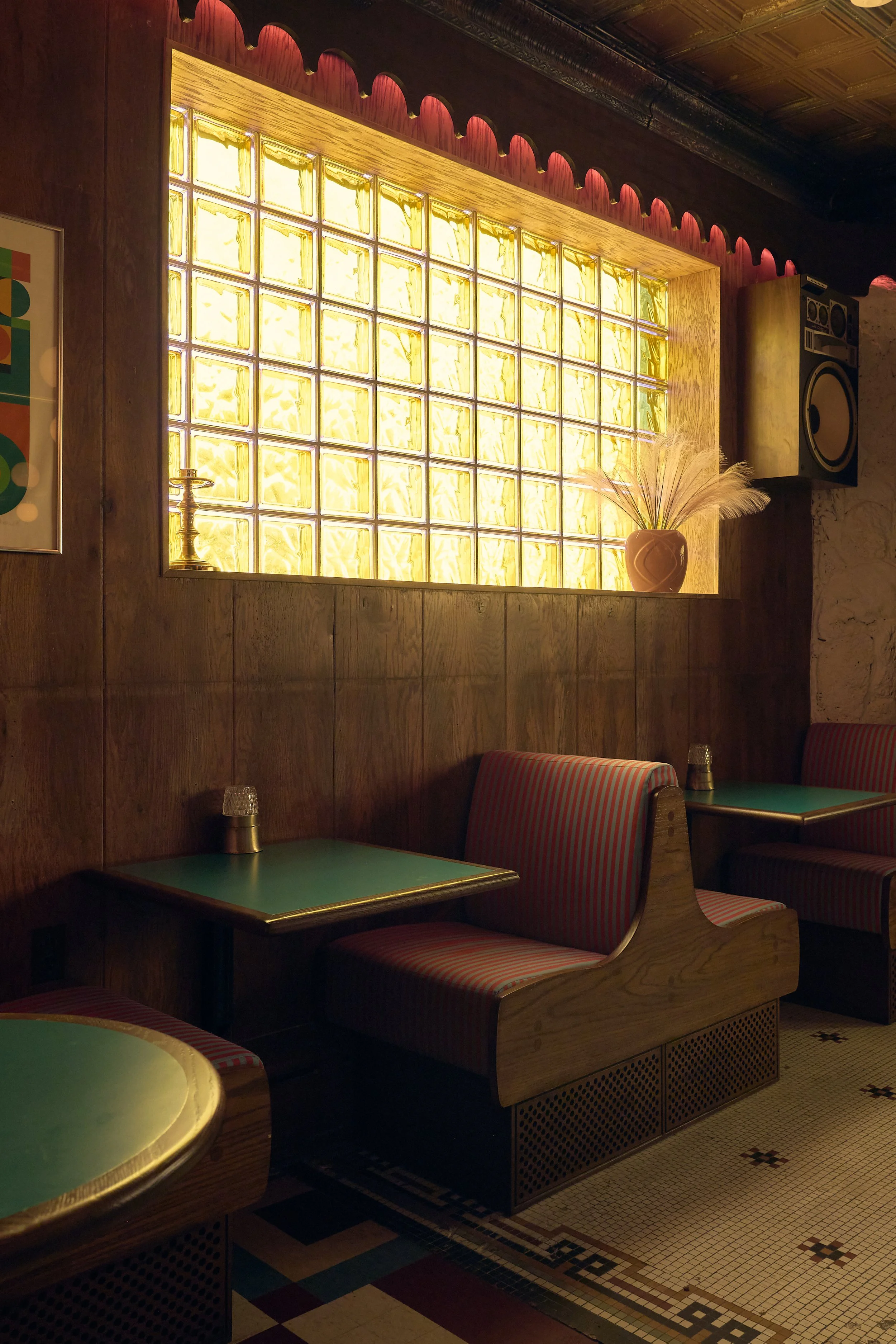 An interior view of a vintage-style restaurant or café, featuring wood-paneled walls, a large window made of glass blocks letting in yellow light, striped upholstered benches, green tables with brass edges, and decorative ceramic vases with dried pampas grass.