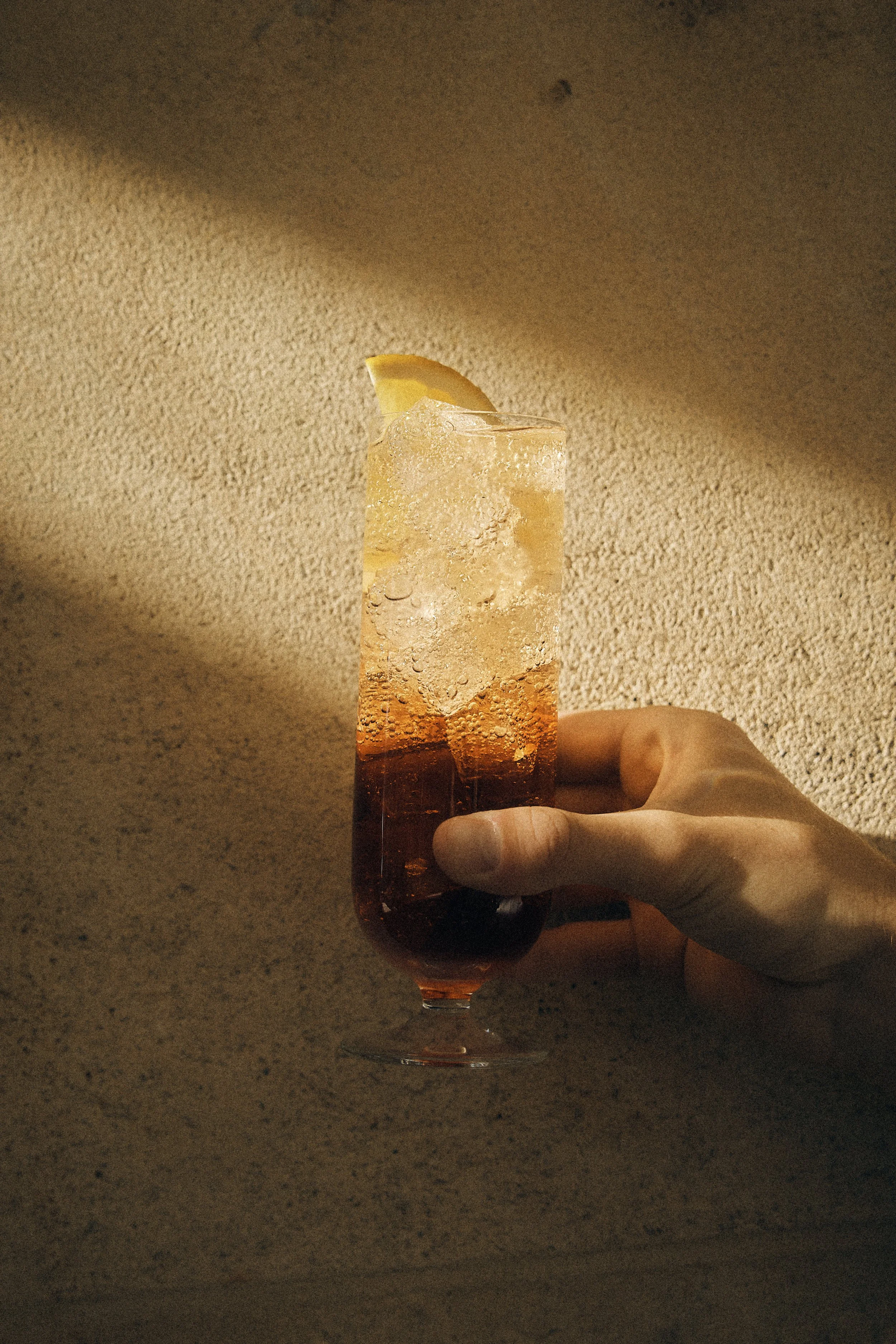 A hand holding a tall glass of sparkling beverage with ice and a lemon wedge garnish.