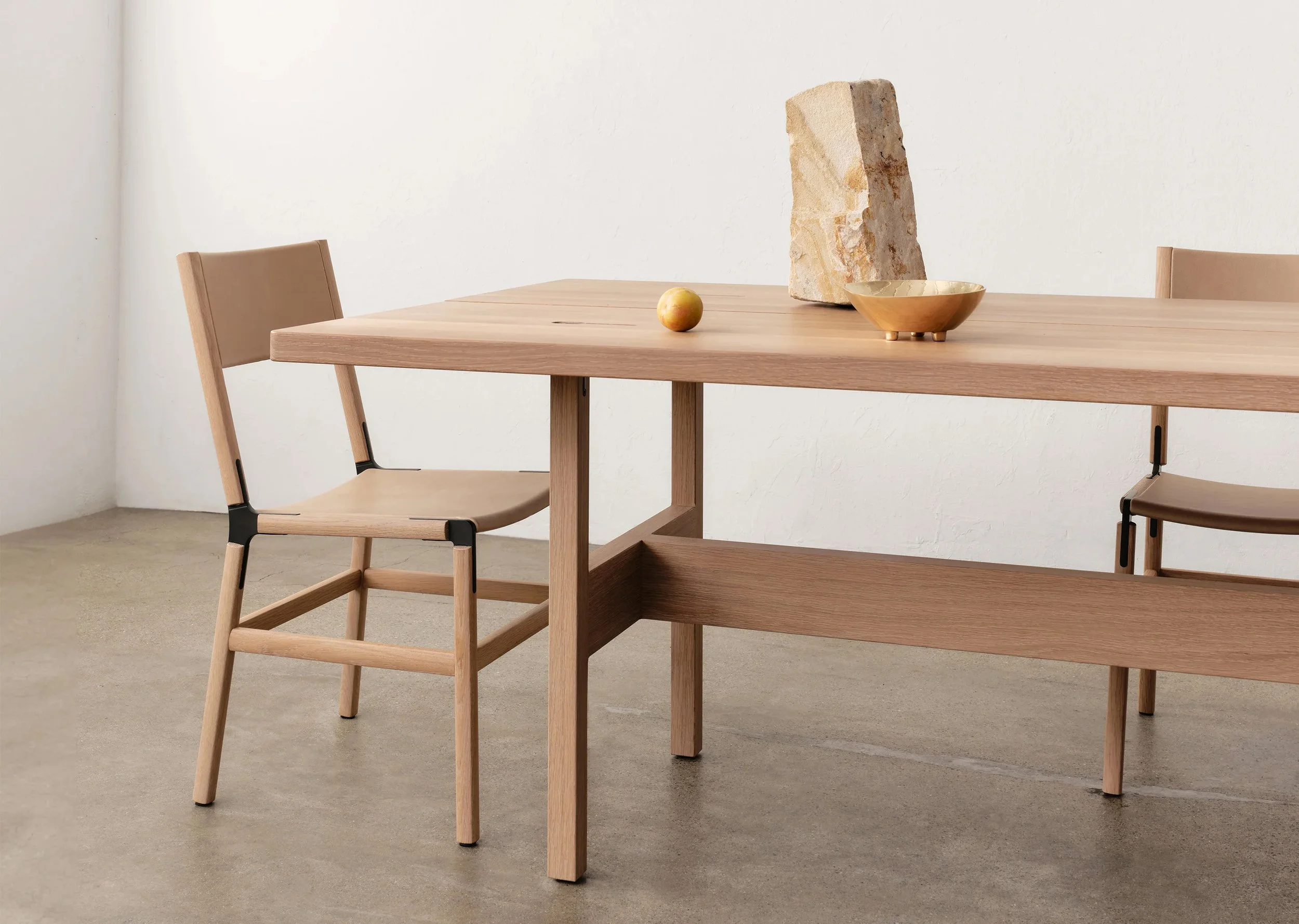 A modern wooden dining table with two matching chairs, set against a plain white wall. The table has a decorative stone slab, a small golden bowl, and a peach placed on it.