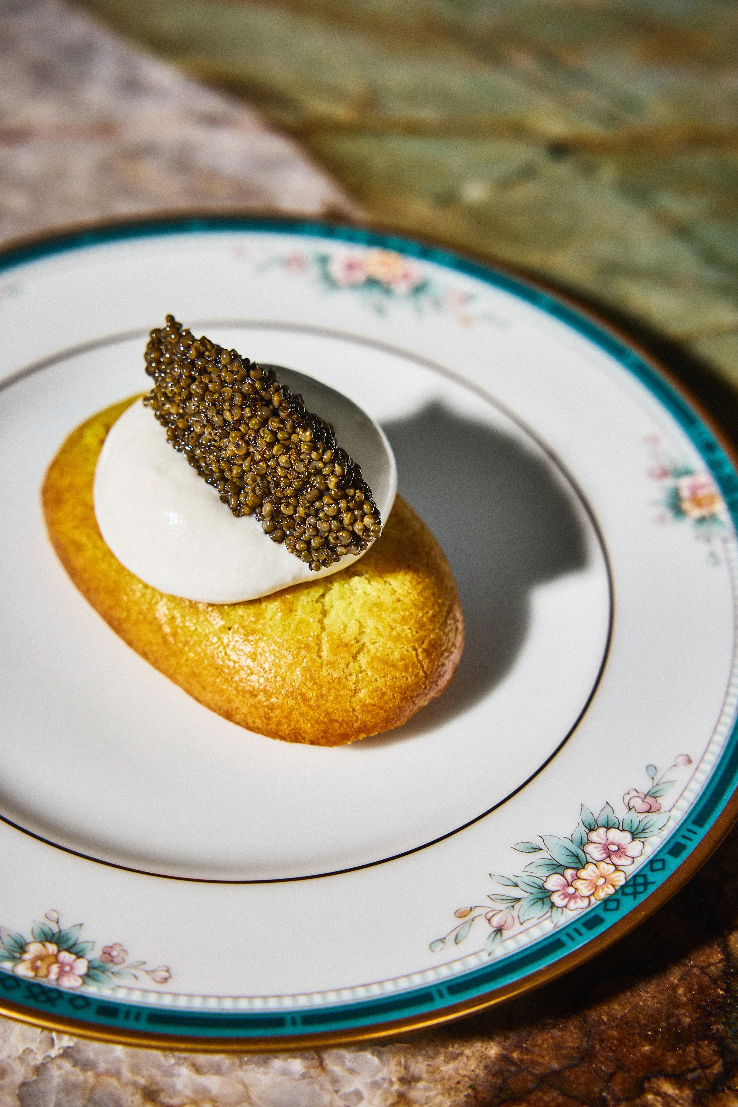 A dessert with a golden baked cupcake topped with a dollop of white cream and a black caviar garnish, served on a decorative floral patterned plate.