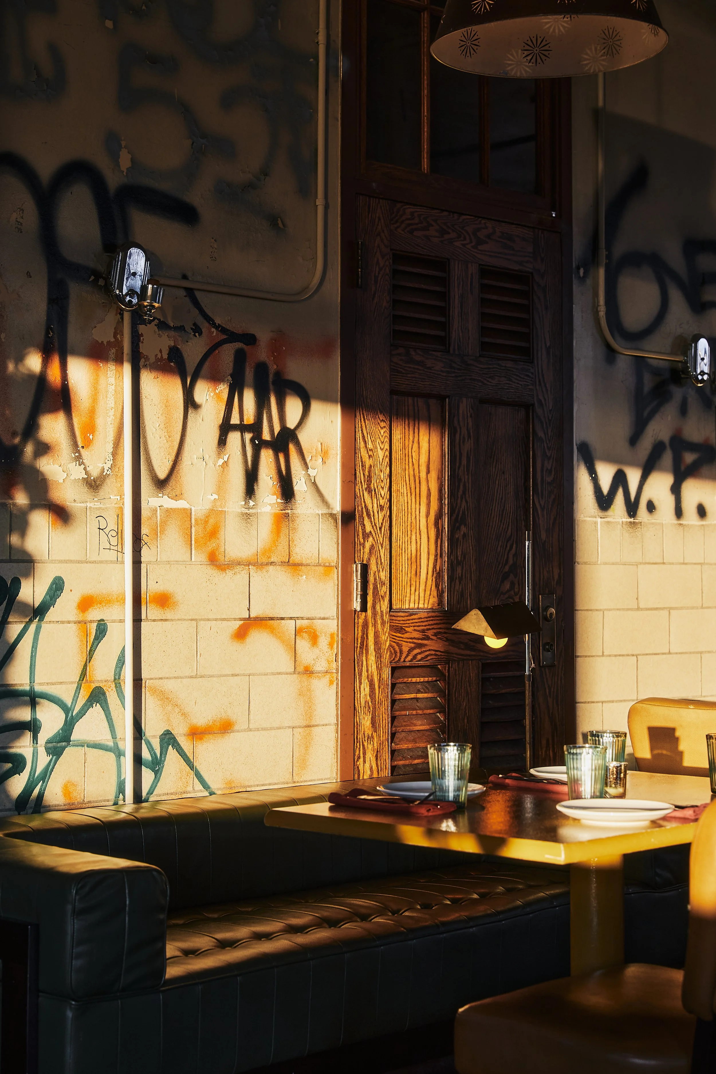 A cozy restaurant interior with a black leather tufted bench, wooden table with plates, glasses, and napkins, a wall with graffiti art, hardwood door, and pendant light fixtures overhead, illuminated by warm lighting.