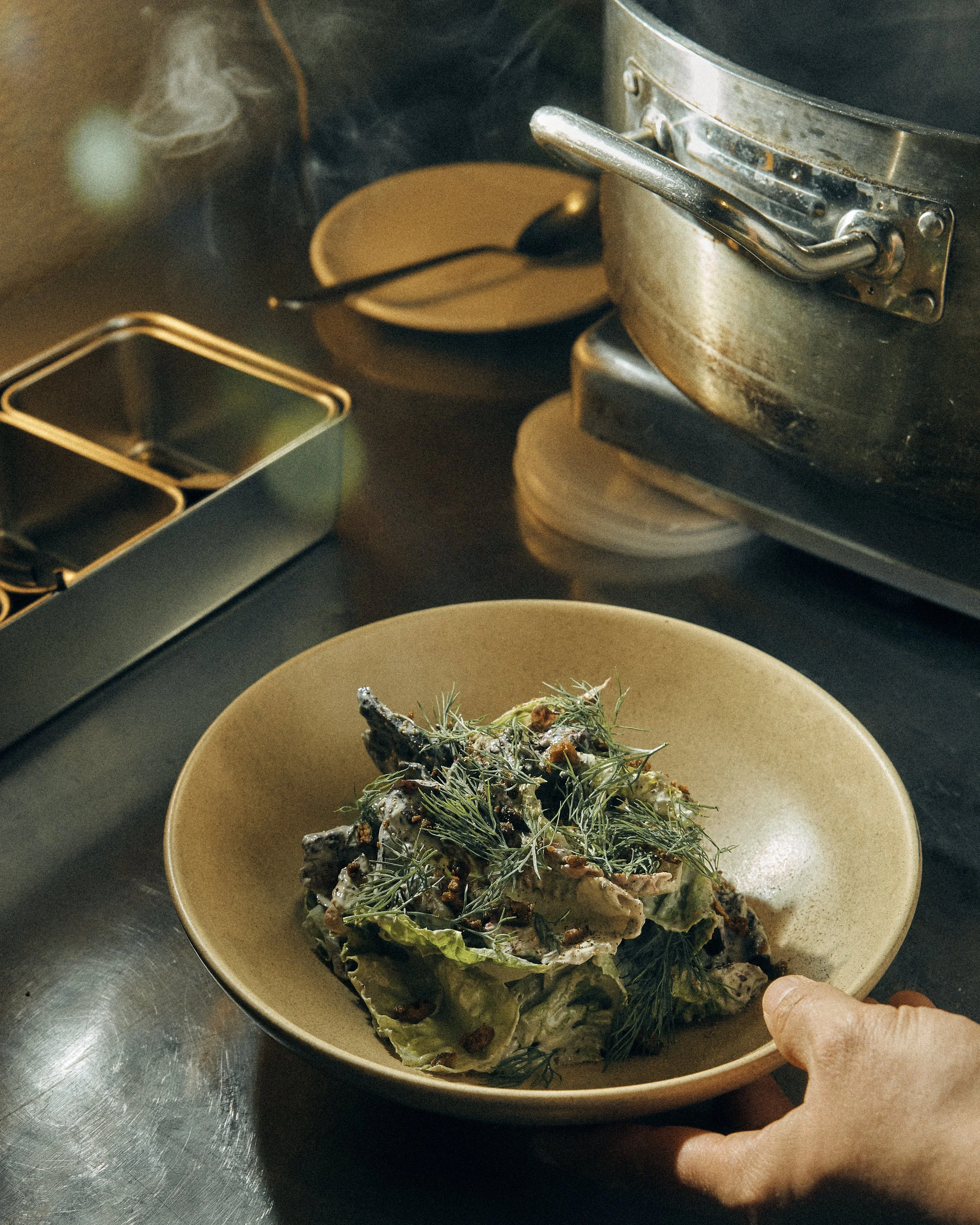 A person's hand is reaching into a beige bowl containing a cooked dish garnished with herbs, placed on a dark countertop. The background shows a stove with a pot, a spoon in a bowl, and some metal containers.