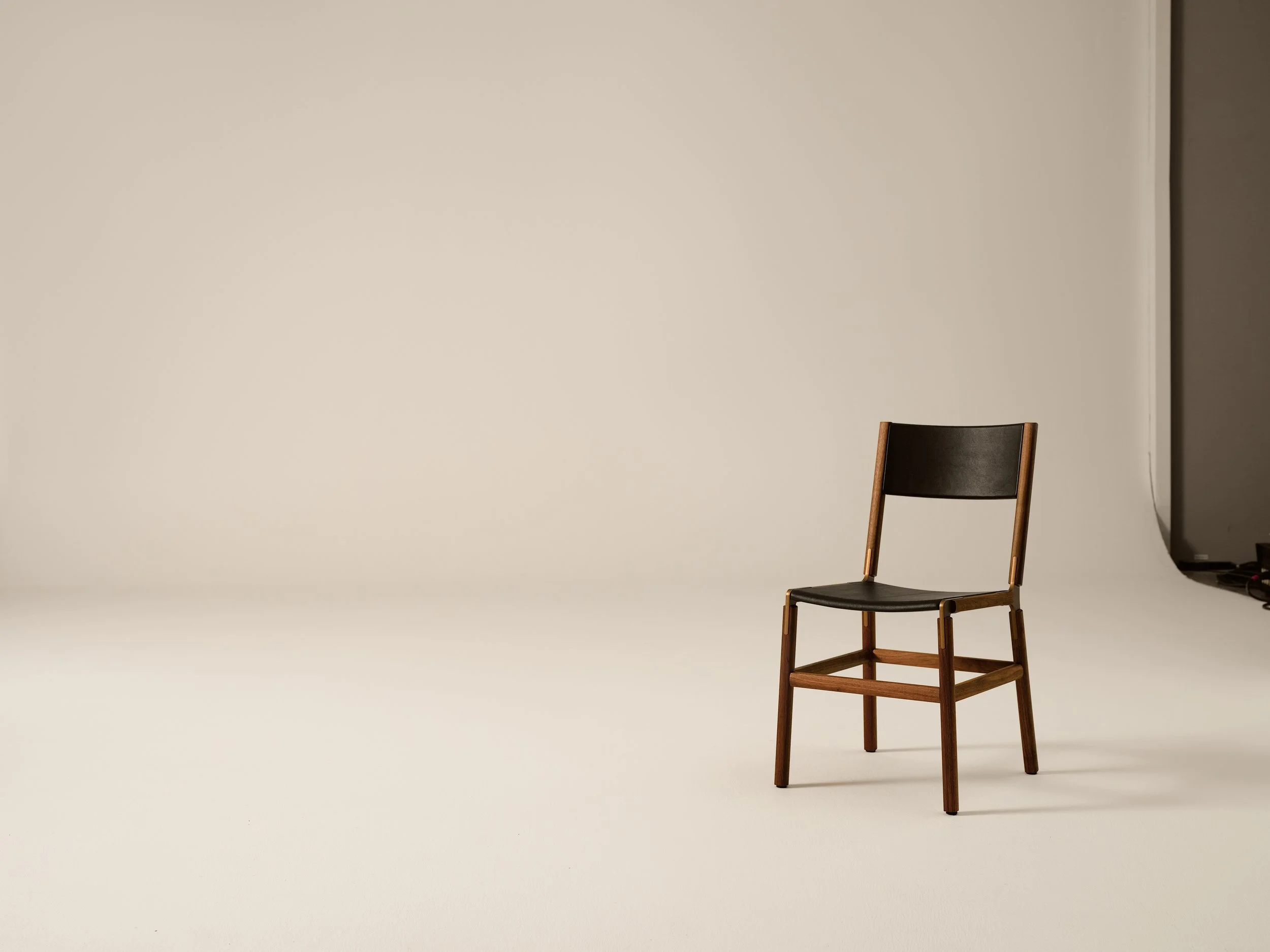 A black chair with wooden legs and a wooden frame, positioned against a plain beige backdrop, with part of a hallway and a gray appliance visible on the right side.