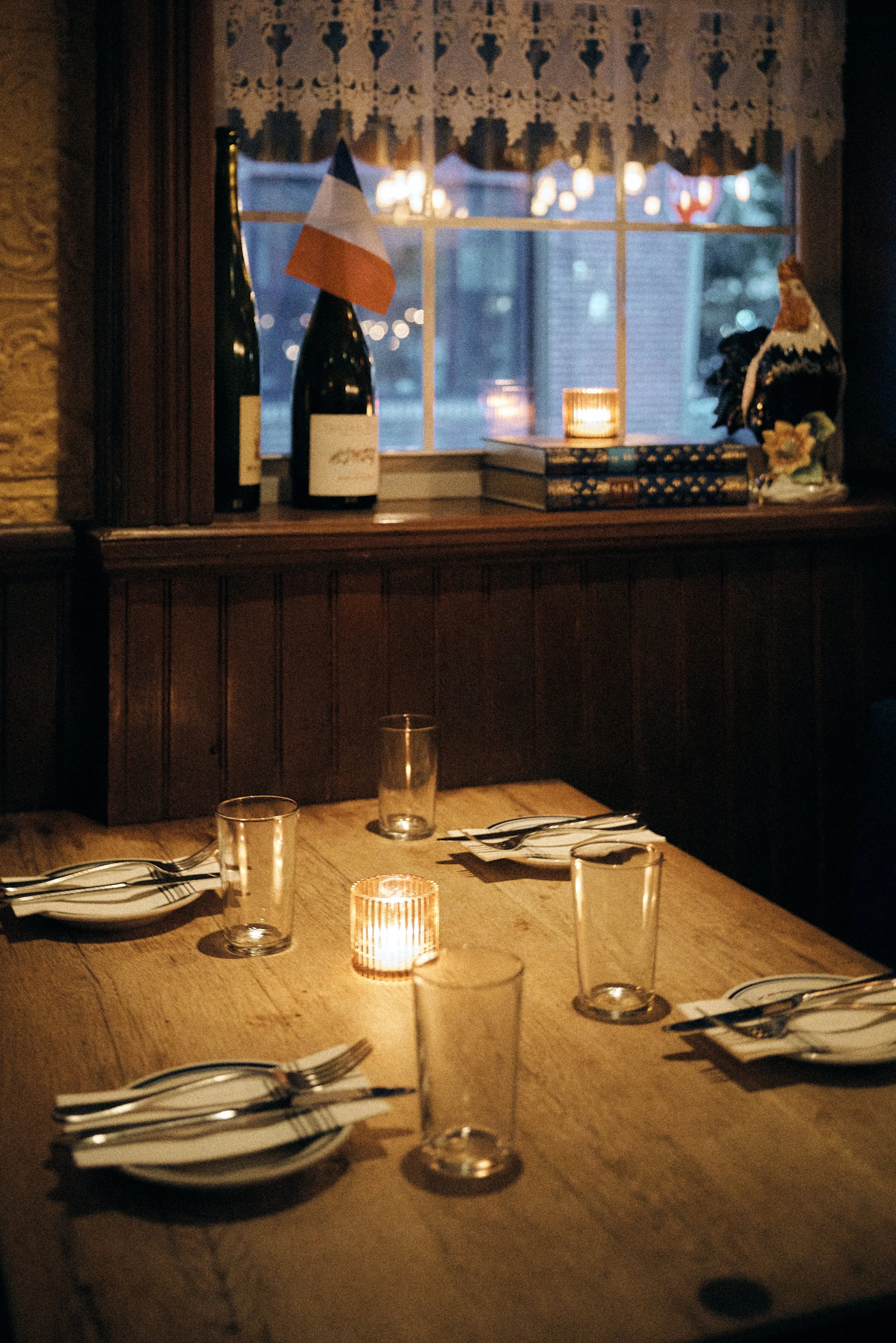 A cozy dining room scene featuring a wooden table set with plates, forks, and glasses. A small lit candle is on the table, and a window with lace curtains shows a blurred outdoor view. On the windowsill, there are two bottles, a small stacked set of books, a candle, and decorative items including a ceramic rooster and a small floral arrangement. The lighting is warm and inviting, suggesting an intimate dinner setting.