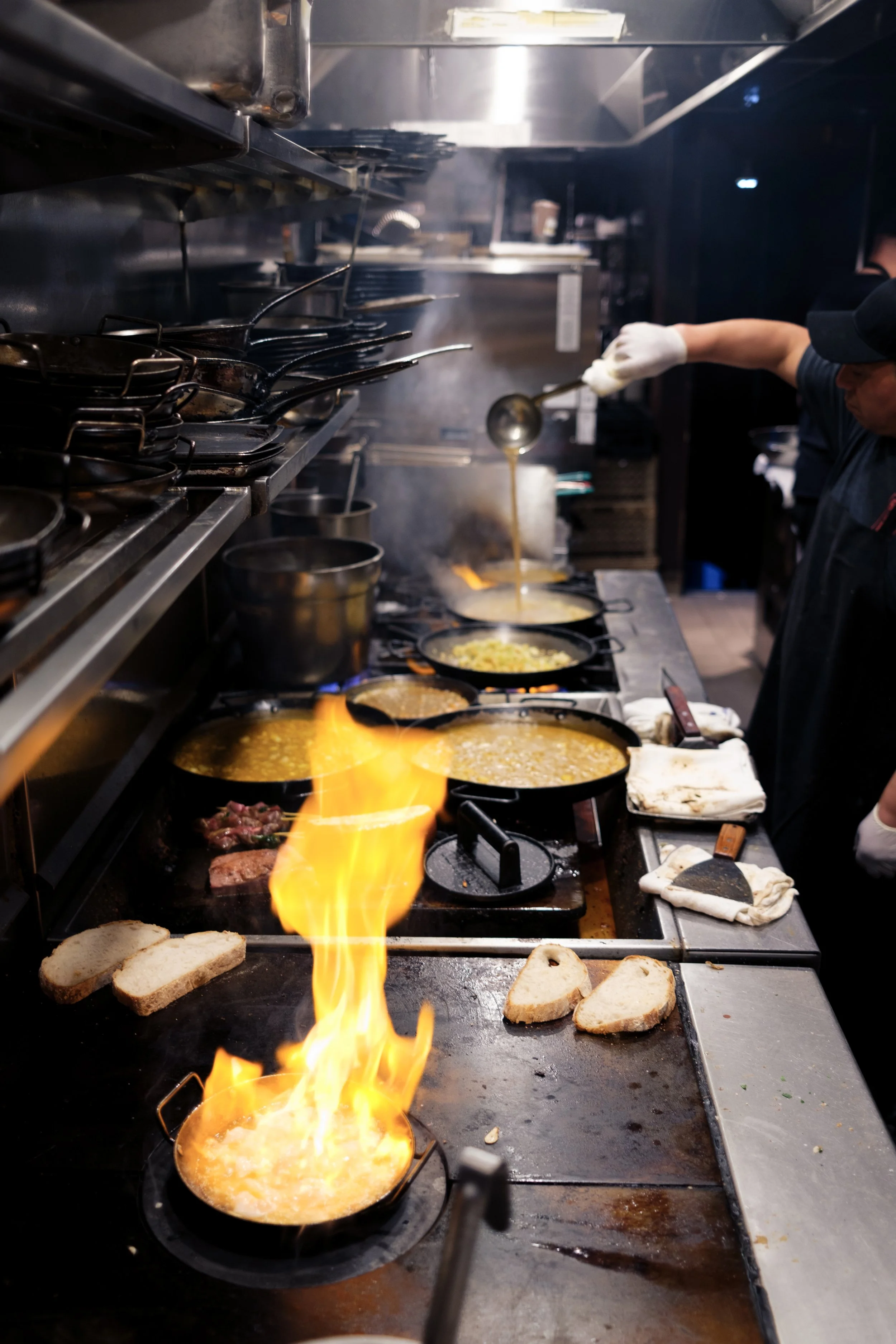 A professional kitchen with multiple pans on a stove, cooking various dishes. Flames are visible under one pan, and a chef is adding ingredients from a ladle. Slices of bread are on the counter.