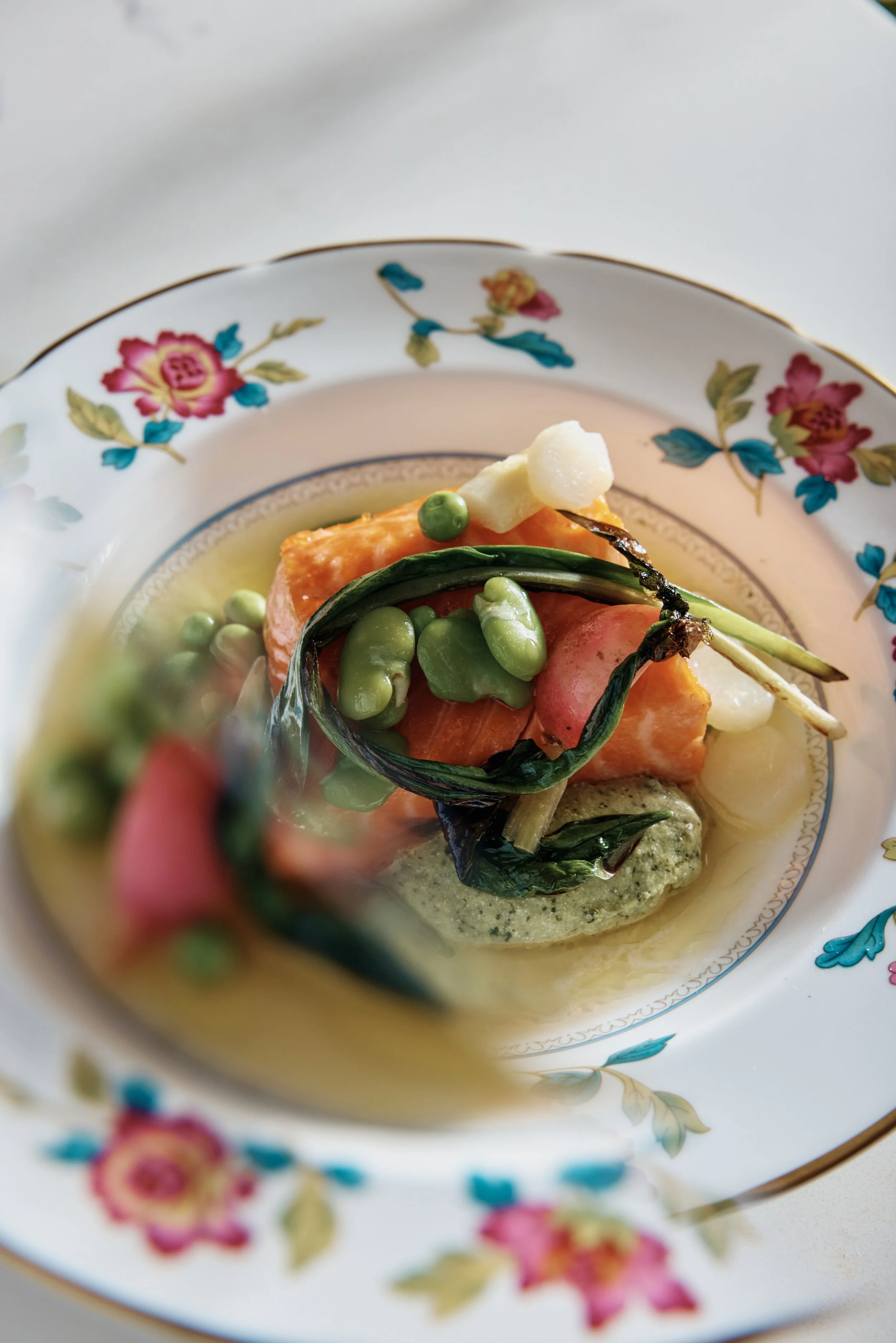 Close-up of a decorative plate with floral patterns, containing cooked fish, green peas, and mashed potatoes, with sauce or puree underneath.