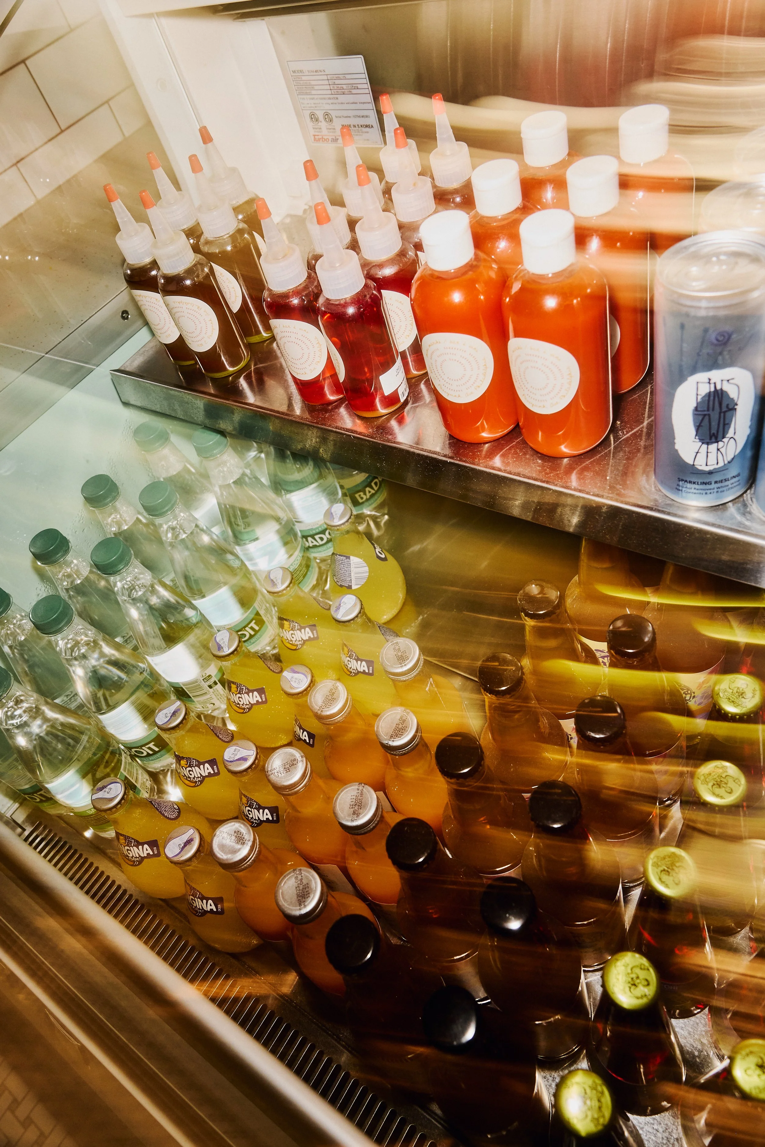 Various bottles of colorful beverages inside a refrigerated display case, including bottles with orange, red, and clear liquid, some labeled with brand names like Angie and S. Zero.