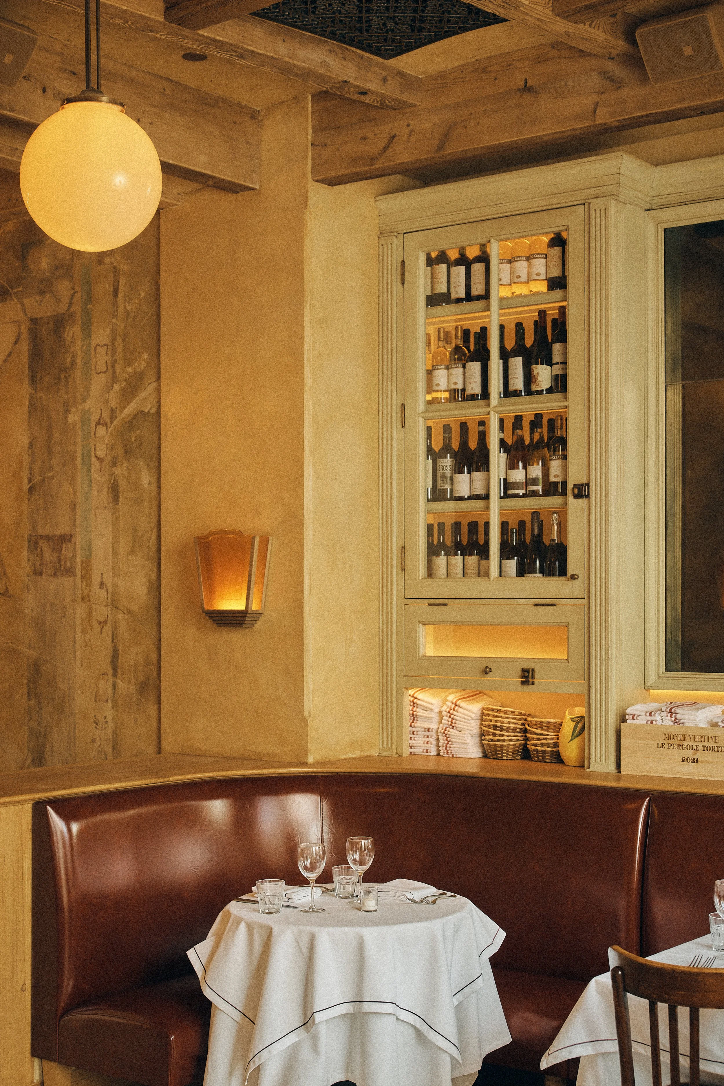 A cozy restaurant interior features a dining table set with wine glasses and silverware, a white tablecloth, and brown leather booth seating. The wall includes vintage-style lighting and a display cabinet filled with wine bottles, with a rustic wall to the left and wooden beams on the ceiling.
