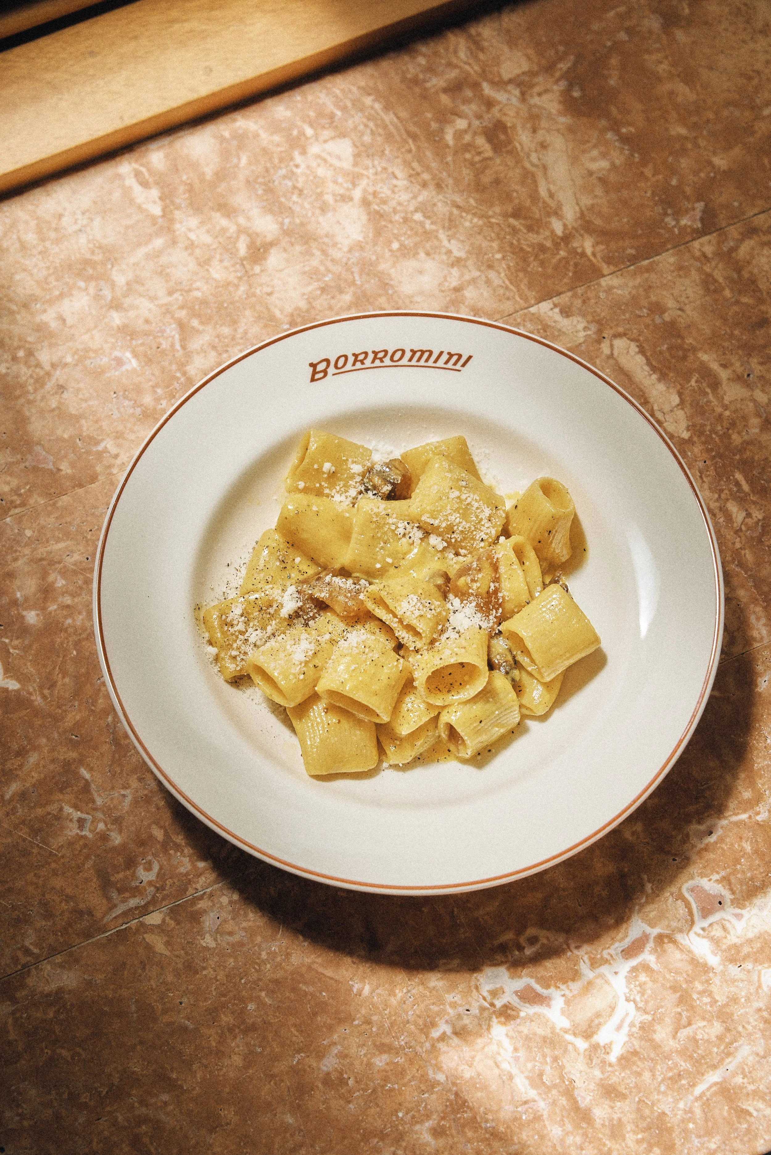 A white plate with the word 'BORROMINI' on the rim, containing cooked pasta with cheese and possibly mushroom pieces, placed on a brown marble countertop.