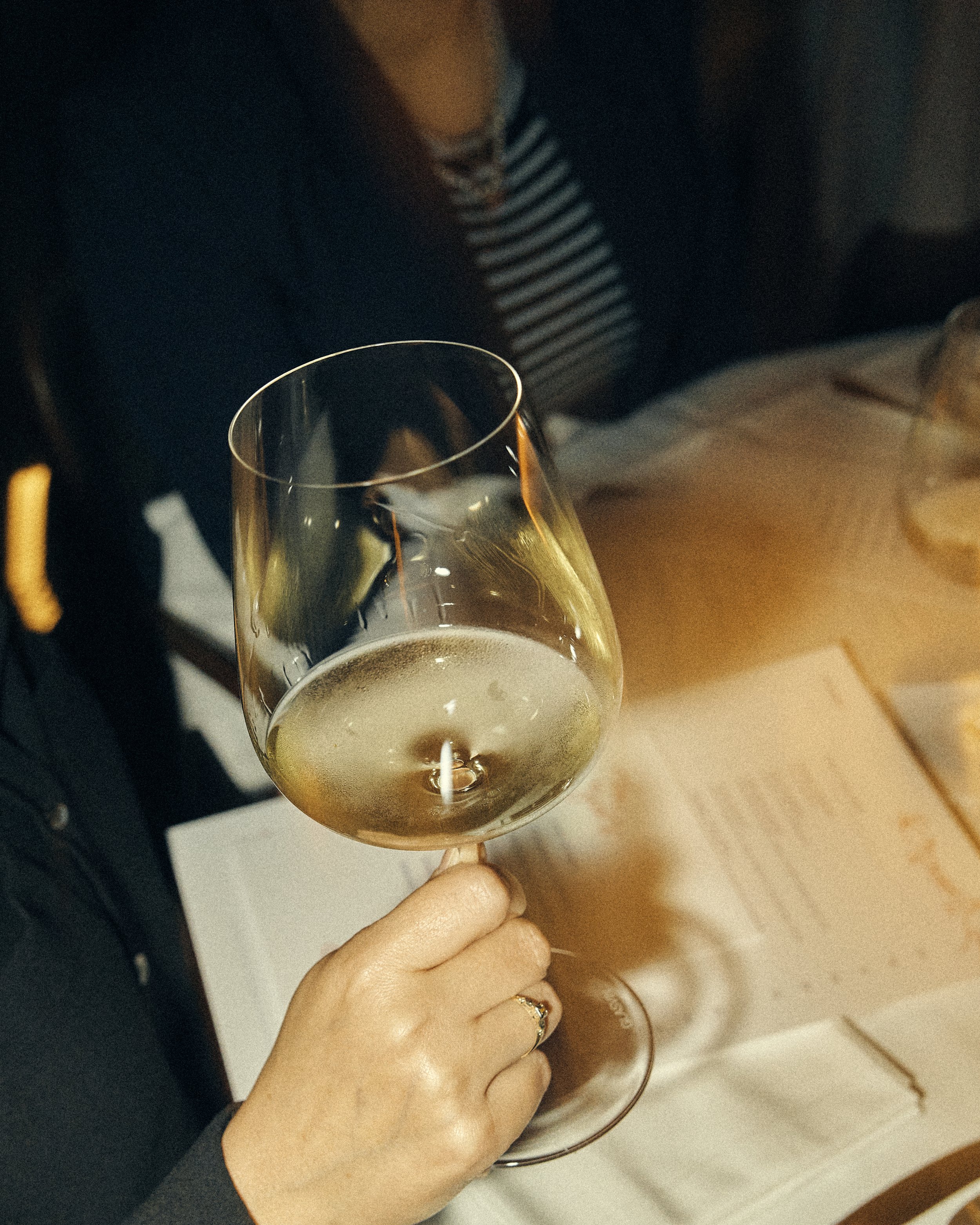 Person holding a glass of white wine, wearing a black jacket and a striped shirt, in an indoor setting.