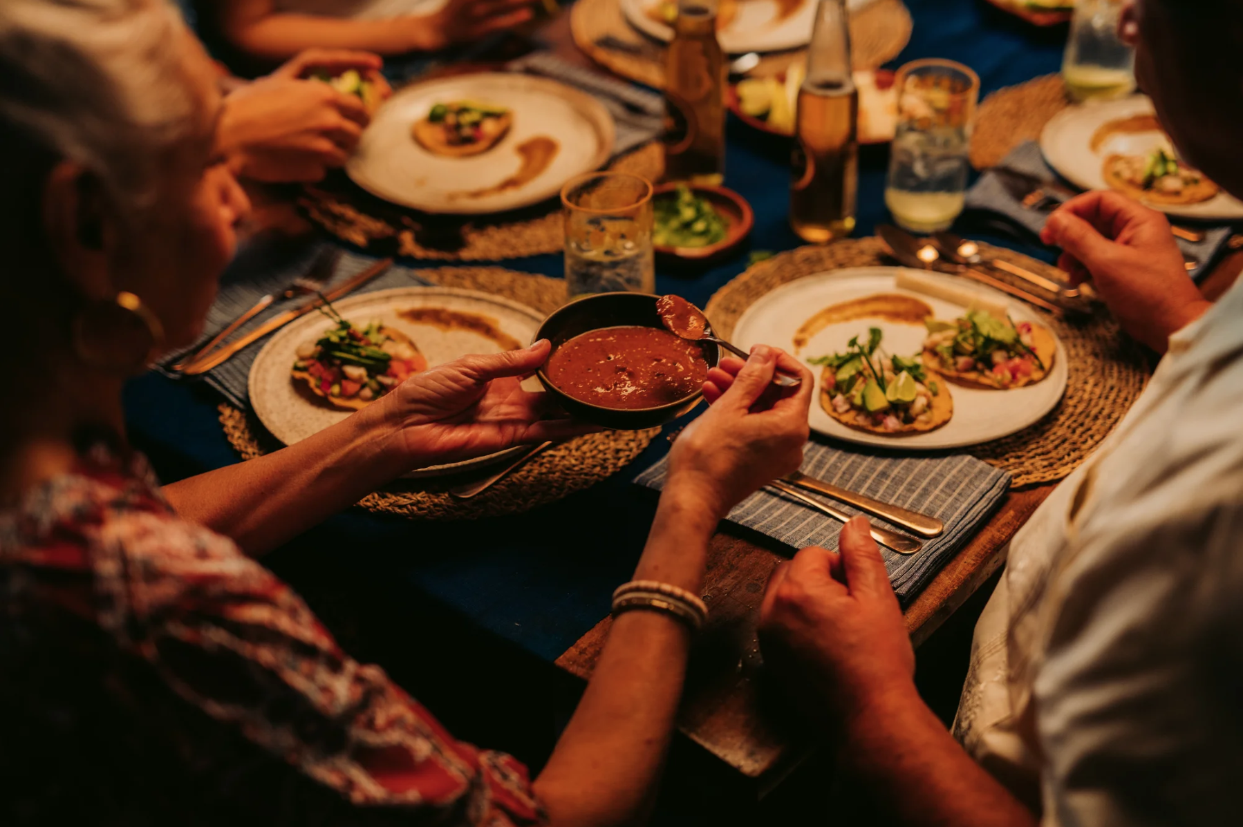 A group of people gathered around a dining table enjoying a meal with plates of tacos, drinks, and a bowl of sauce, with one person serving sauce onto a taco.