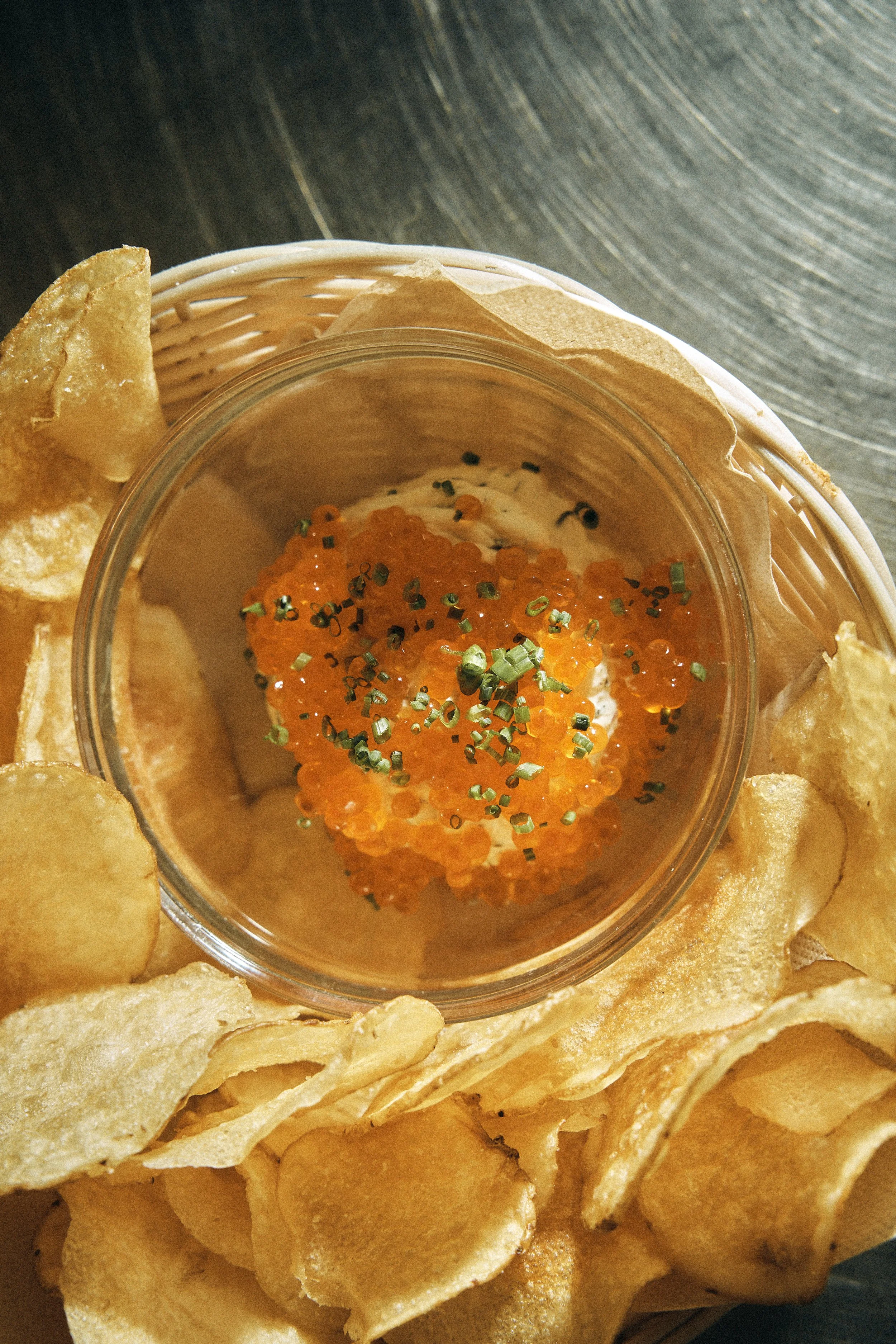 Close-up of crispy chips surrounding a small container of dip topped with orange fish roe and green seasoning.