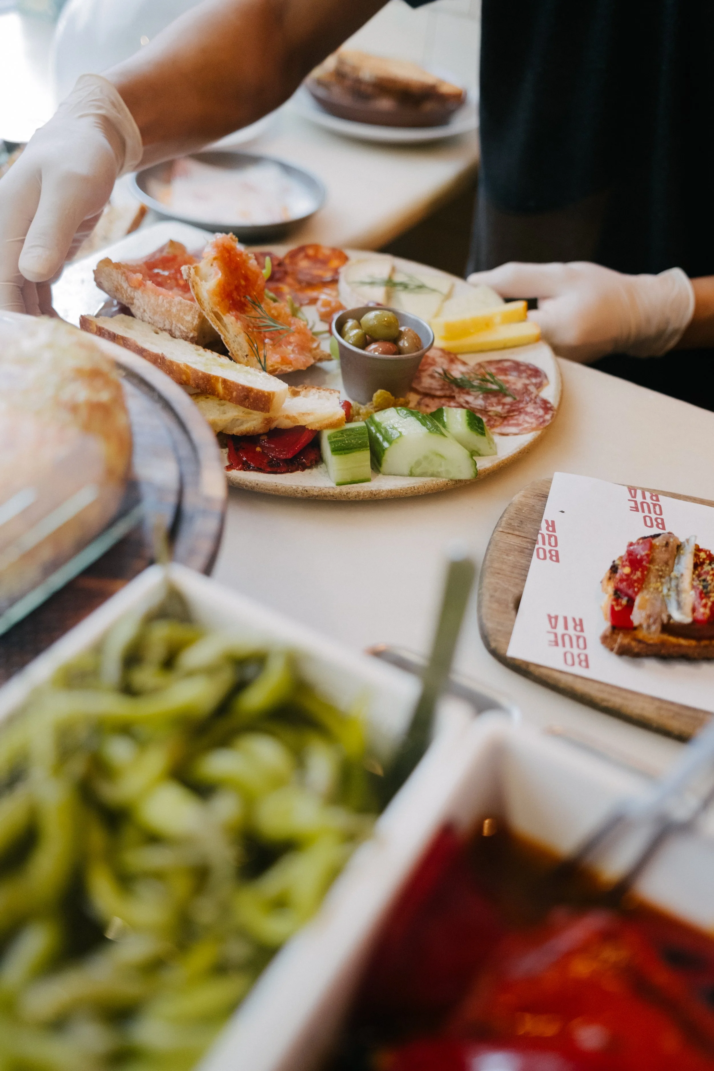 A plate of assorted appetizers, including olive olives, sliced cucumbers, slices of cured meats, bread slices with toppings, and grilled vegetables, with some dishes in the background.