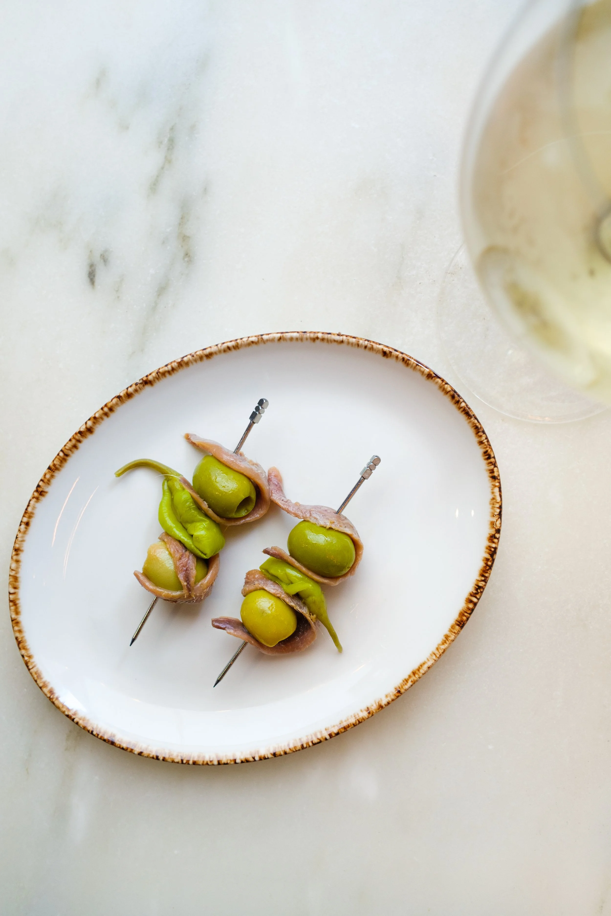 Three skewers with green grapes wrapped in prosciutto on a white and brown ceramic plate, placed on a marble surface, with a bowl of creamy dip or sauce in the background.