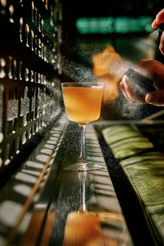 A bartender spray garnishing salt onto a cocktail in a glass with a lime wedge on the side, with a dark background.