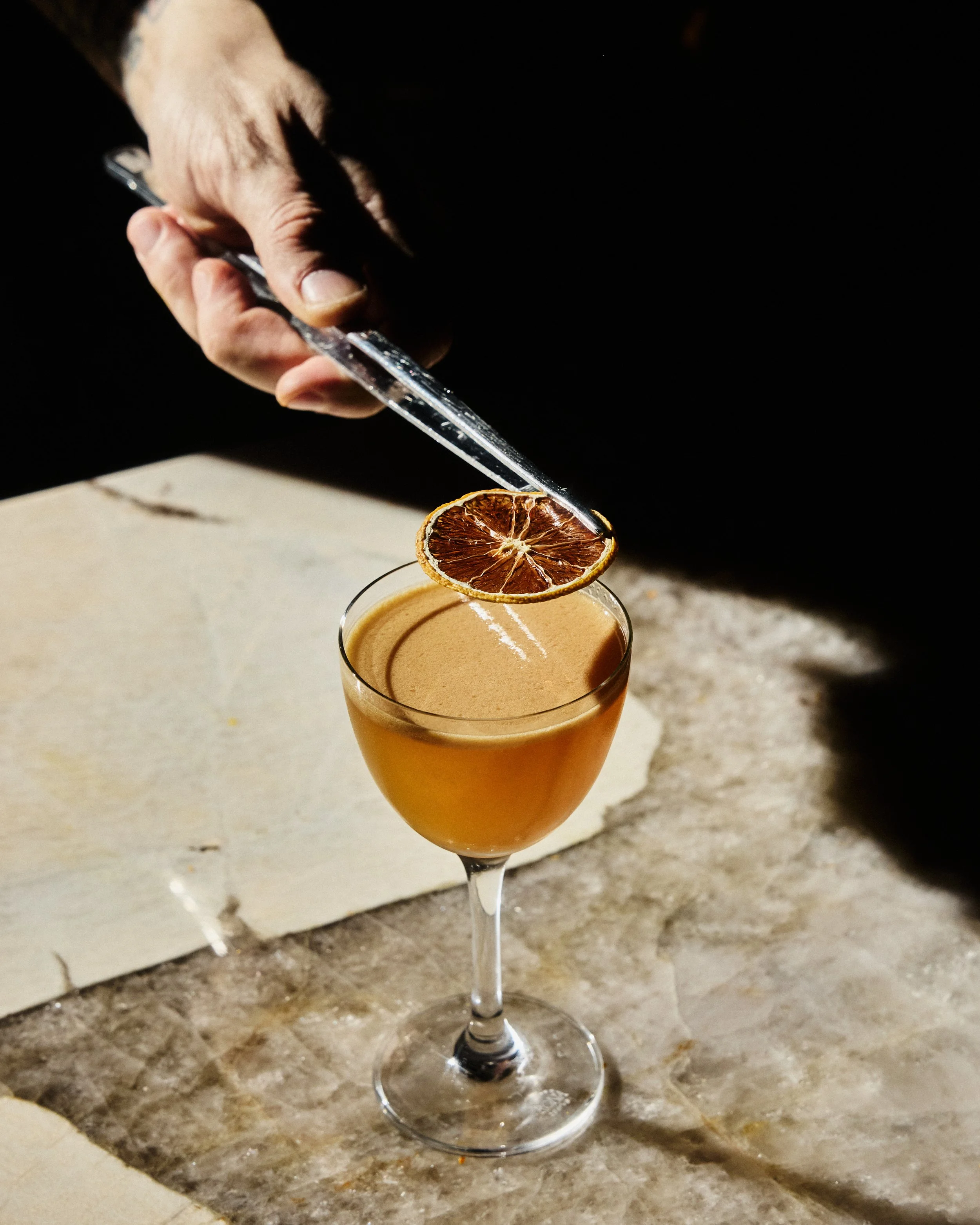 A glass of brown cocktail garnished with a slice of dried blood orange, placed on a marble surface with a dark background.